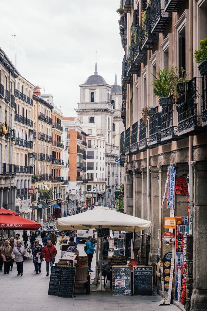 Bustling European street with people walking, outdoor cafés, menu boards, and tall buildings on both sides—a familiar sight for students in Madrid seeking lively atmospheres. In the background, a church with a dome and bell tower rises under a cloudy sky, inviting visitors to explore more Madrid activities in this vibrant city.