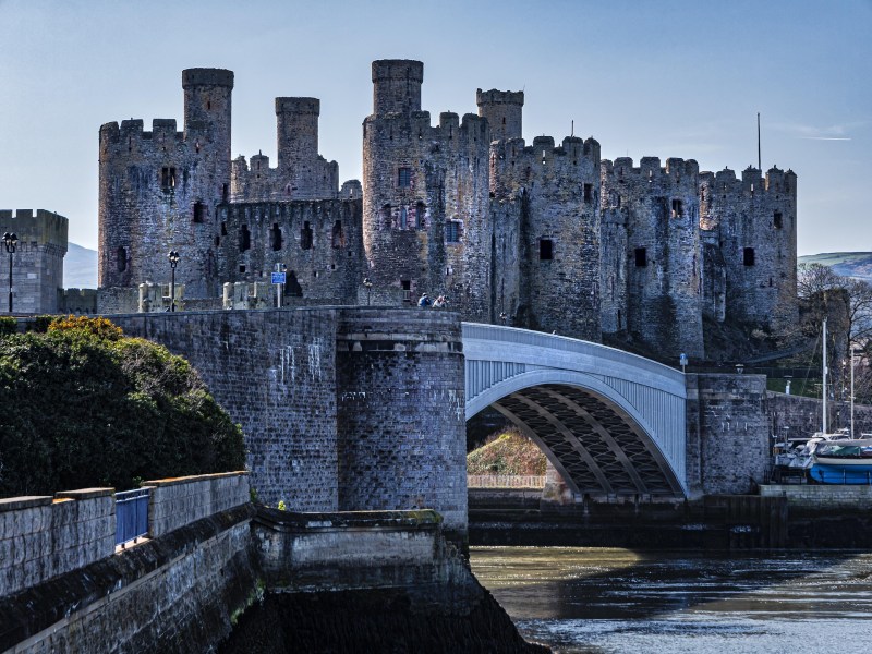 A large stone castle with multiple towers stands behind an arched bridge over a river on a clear day. The castle’s walls and turrets are weathered, and a few people—perhaps students on day trips from Manchester—are visible on the bridge.