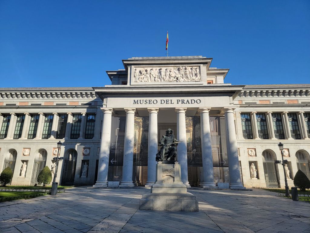 The Museo del Prado building, with its tall columns, a statue in front, and a clear blue sky above, stands as an iconic destination for students in Madrid. A flag flies atop the roof, making it a must-see landmark among the many things to do in Madrid.