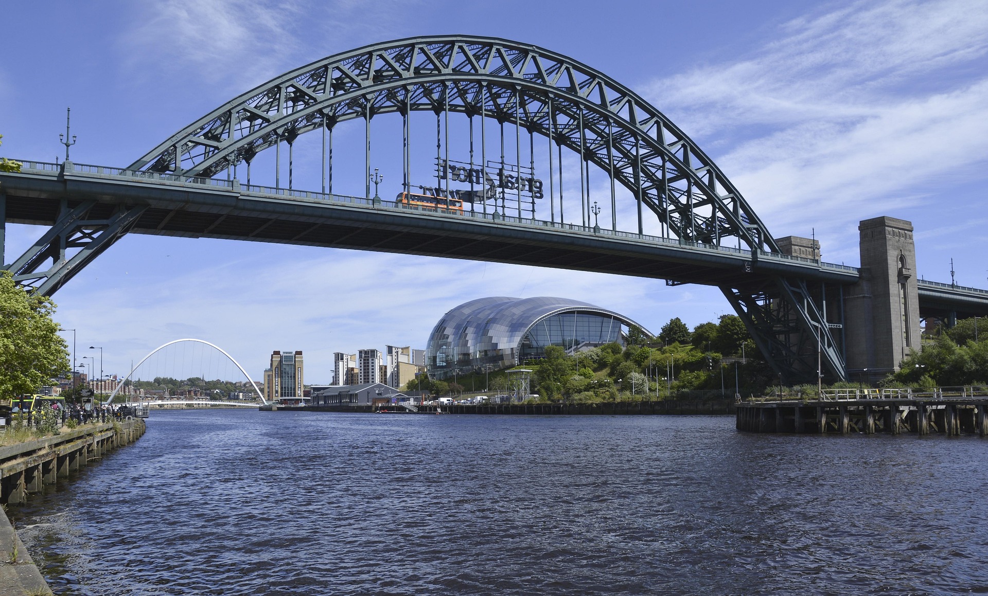 A large steel arch bridge spans over a river, with a modern glass building and another arched footbridge in the background under a blue sky. Trees and buildings, including some of the best places to live Newcastle has to offer for students, line the riverbanks.