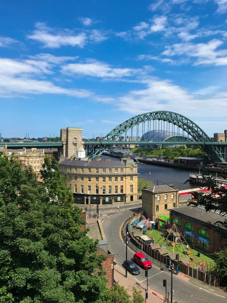 View of Newcastle with a large green arch bridge over a river, surrounded by buildings, a playground, and a red car on the road. This vibrant cityscape under a blue sky with scattered clouds makes Newcastle one of the most appealing places to live—especially for students drawn to its lively atmosphere and scenic beauty.