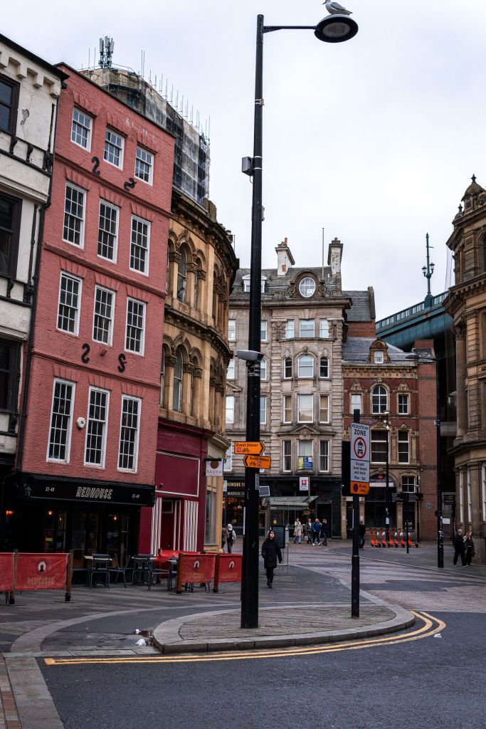 Historic city street with a red, slanted building on the left, surrounded by older architecture—showcasing one of the best places Newcastle has to offer. Several street signs and a few people are visible under a cloudy sky.