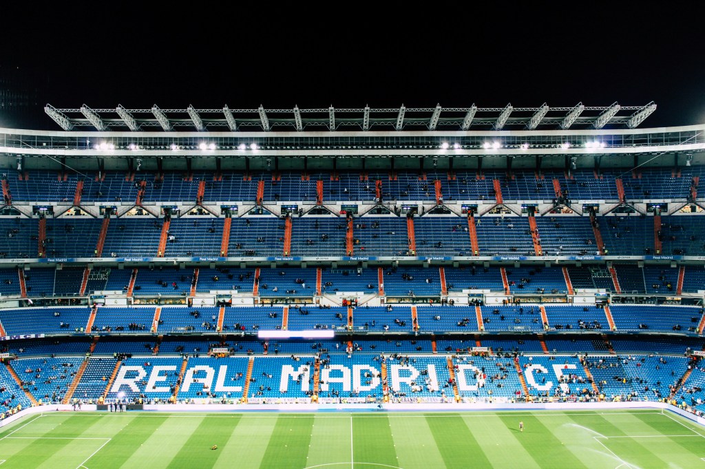 A brightly lit, empty football stadium with blue seats spelling "REAL MADRID C.F." on one side, seen from the stands facing the pitch at night—a must-see for anyone looking for things to do in Madrid or exploring unique student activities Madrid has to offer.