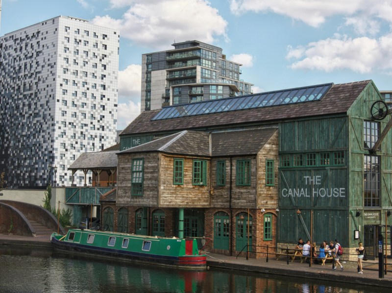 A green and wooden building labeled “The Canal House” sits by a canal with a green boat; modern high-rise buildings and a blue sky with clouds are in the background. Six people walk on the canal path, showcasing why this vibrant area is often counted among the best places for students to live in Birmingham.