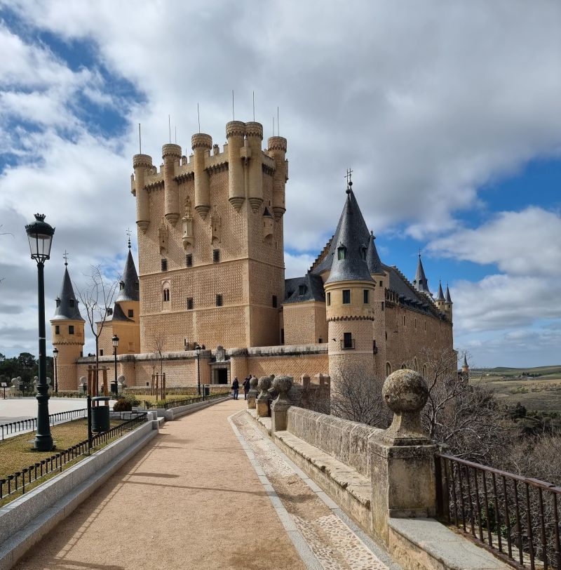 A large, historic castle with round towers and spires sits atop a hill under a partly cloudy sky, next to a pedestrian pathway lined with lampposts and stone railings—a perfect destination for students seeking memorable day trips from Madrid.