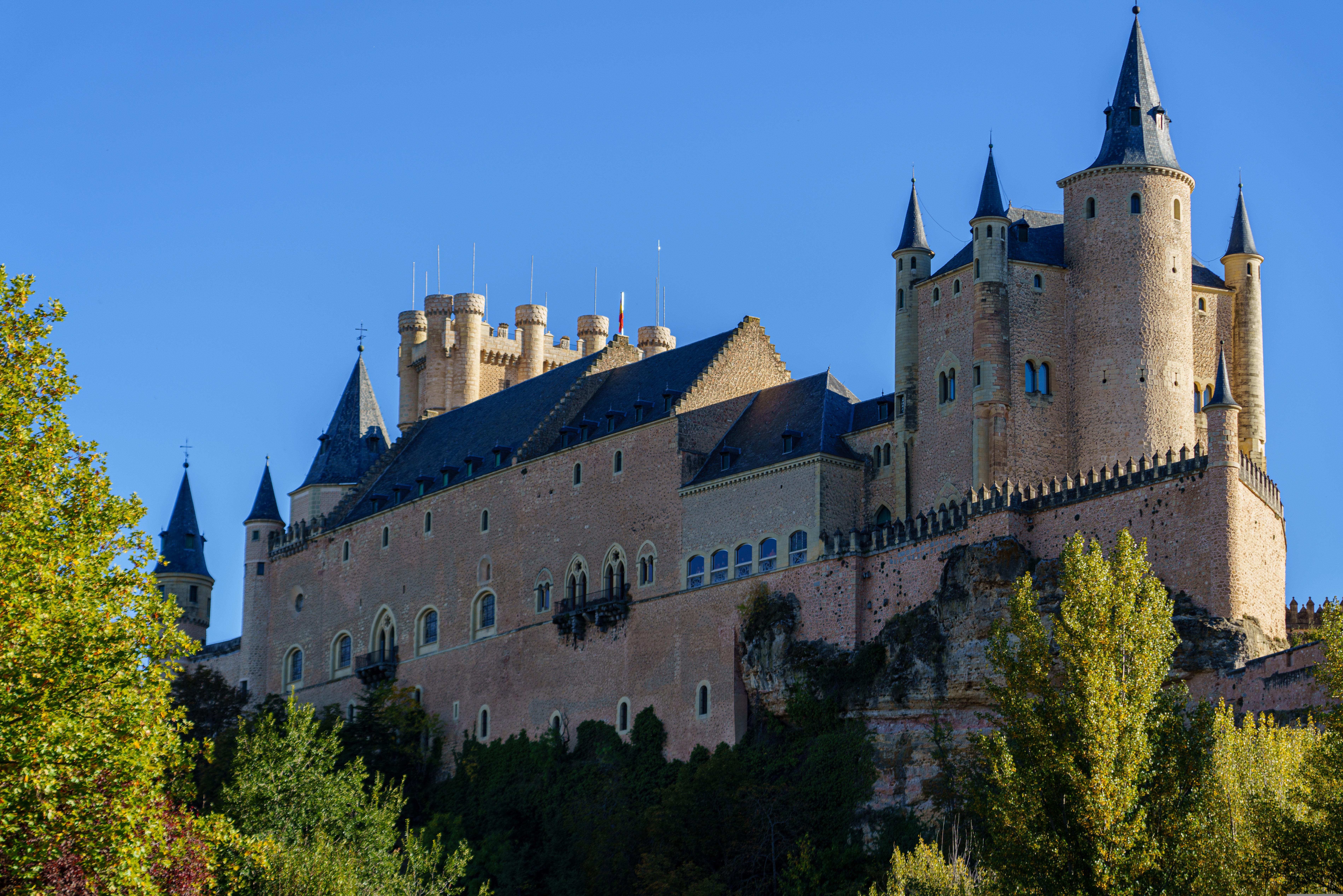 A large stone castle with multiple towers and spires sits on a rocky hilltop, surrounded by green trees under a clear blue sky—an ideal destination for students exploring Madrid or seeking memorable day trips in the region.