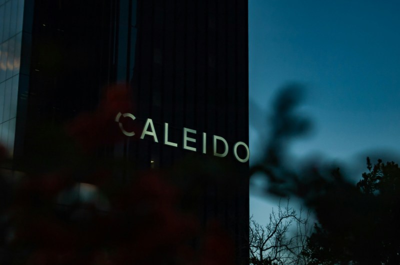 A dark building with illuminated letters spelling “CALEIDO” is partially obscured by blurry branches and leaves in the foreground at dusk, offering a unique backdrop for students near Oria seeking new things to do or engaging activities.