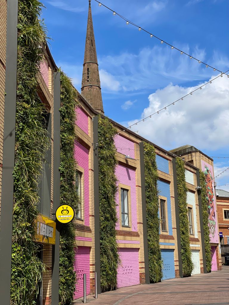 A colorful building with pink, blue, and yellow walls, vertical green plants, and string lights overhead creates a lively atmosphere perfect for students exploring things to do in the area. A church spire rises in the background under a blue sky with clouds.
