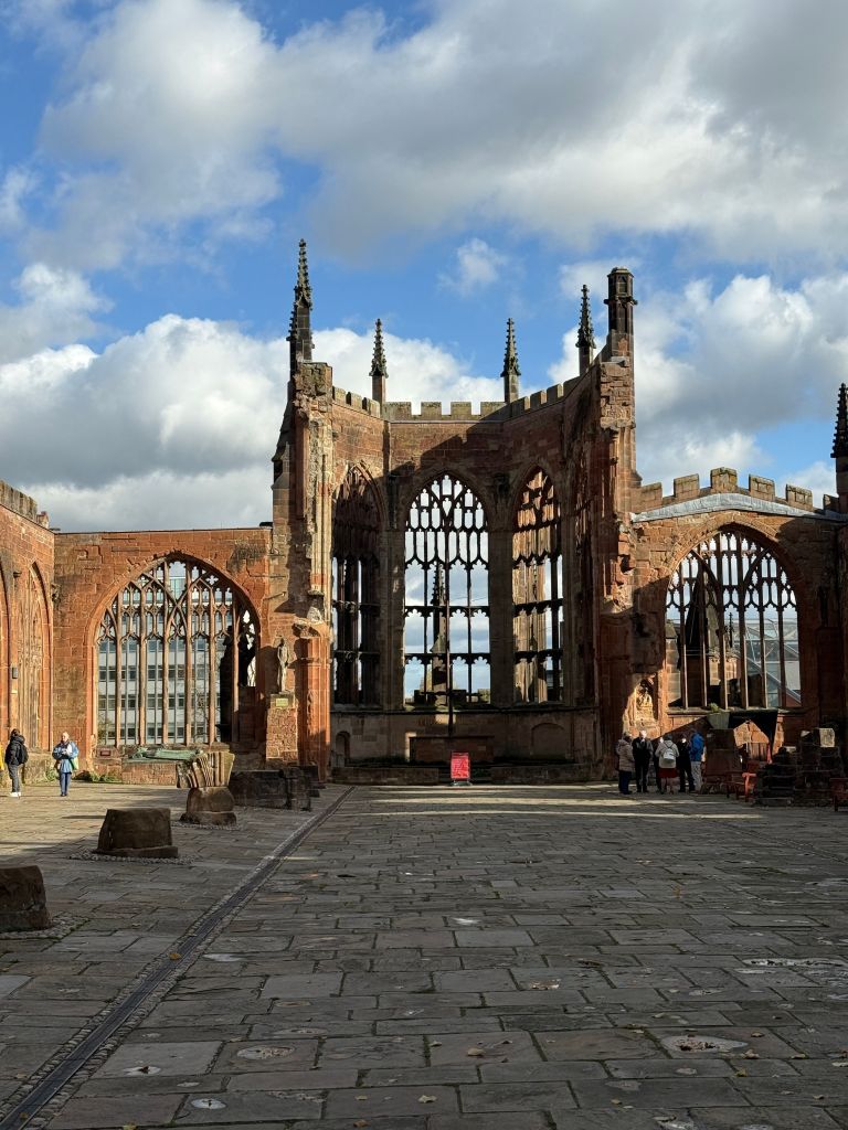 Ruins of a large stone cathedral with tall, arched windows and no roof. A few students walk and stand on the stone courtyard under a partly cloudy sky, discussing things to do after exploring the nearby Copper Towers.