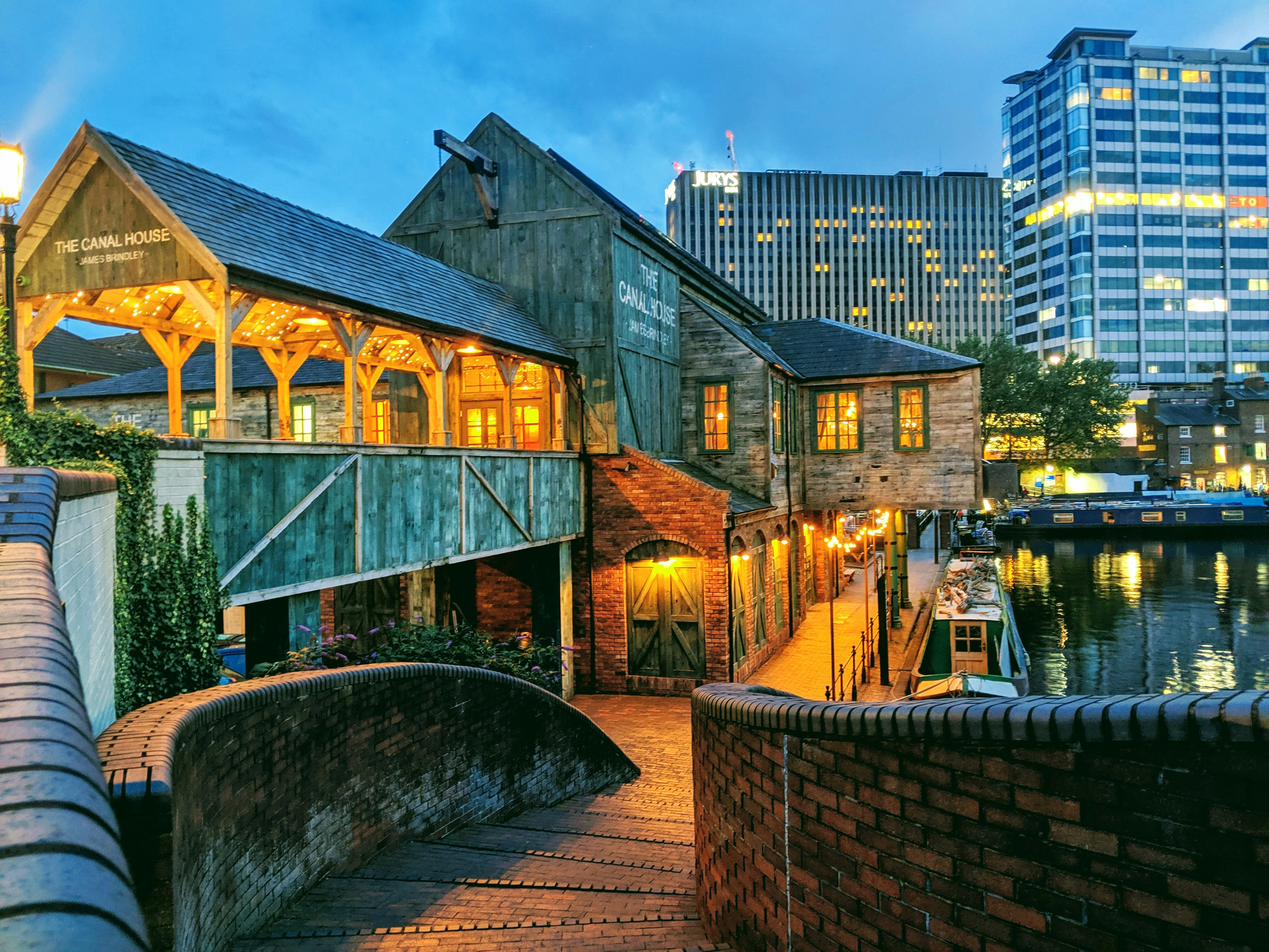A lit riverside restaurant with wooden and brick architecture is beside a canal at dusk. A curved brick walkway leads to the entrance, with city buildings, boats, and nearby student accommodation Birmingham visible in the background—making this area one of the best places to live in Birmingham for students seeking vibrant city life.