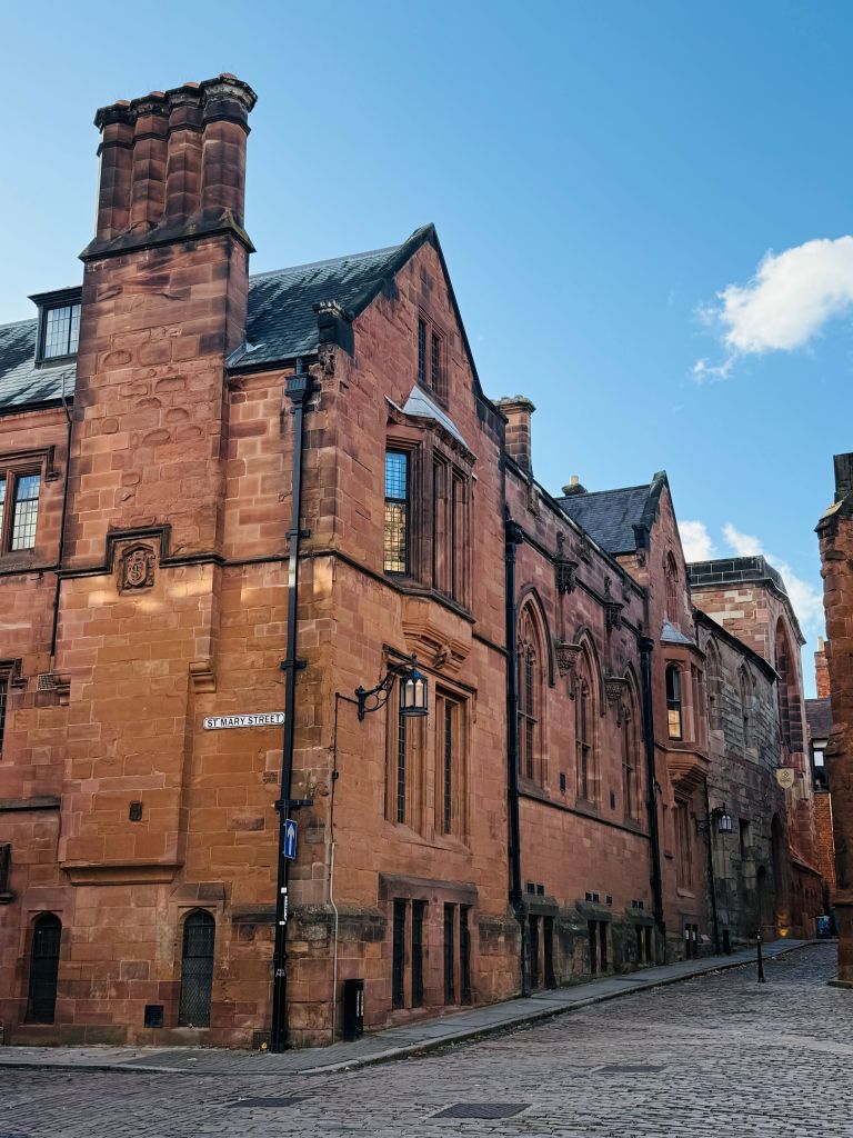 A cobblestone street lined with large, historic red stone buildings under a clear blue sky. Black iron lamps are mounted on the building and a street sign reads “Elphinstone.” Students strolling along the avenue can enjoy discovering things to do nearby, including visiting the iconic Copper Towers just around the corner.
