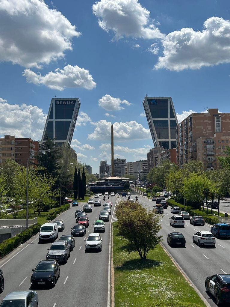 A busy street in the city of Oria, lined with trees and buildings, leads to two leaning towers and a tall monument under a partly cloudy sky, creating an ideal spot for students seeking interesting things to do in the area.
