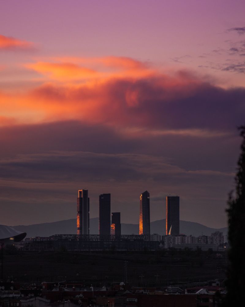 Skyscrapers reflect the orange glow of a sunset under a purple and orange sky, with mountains and a cityscape in the background, creating an inspiring scene for students exploring things to do in Oria.