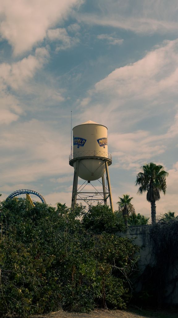 A yellow water tower with the word “KNOWLAND” stands above green trees and palm trees under a partly cloudy sky, with part of a blue roller coaster visible on the left—an inviting spot for students seeking memorable day trips reminiscent of those popular in places like Madrid.