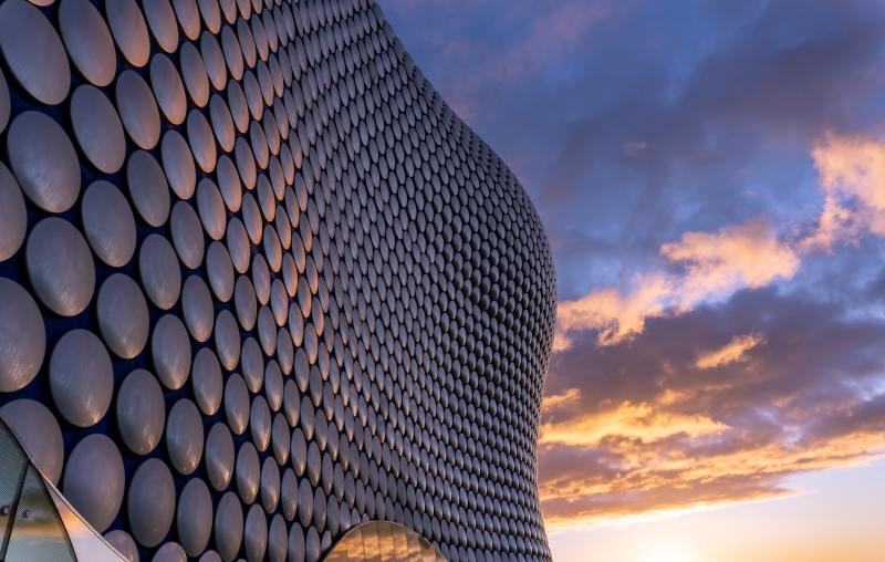 Curved building facade with circular metallic panels reflecting sunlight, set against a colorful sunset sky with scattered clouds—an architectural highlight often found near the best places for students to live in Birmingham.