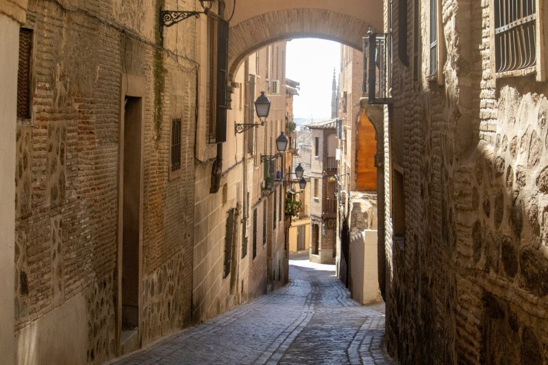A narrow, cobblestone street lined with old brick buildings curves gently under an archway in Madrid, with lanterns hanging on the walls and sunlight illuminating the path ahead—a perfect scene for students exploring the city on day trips.