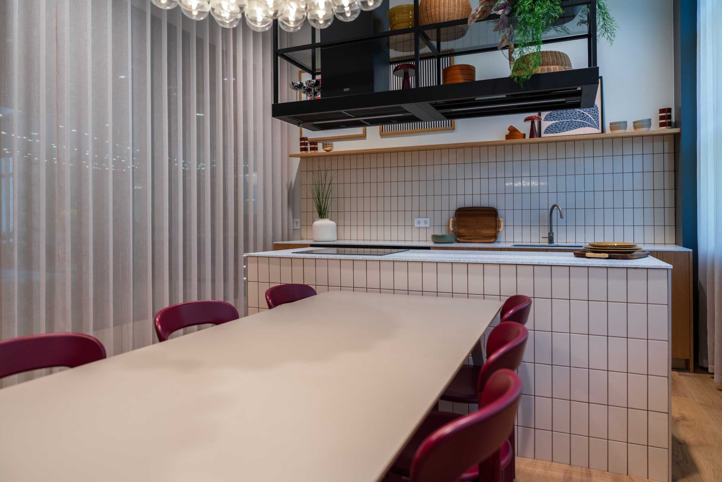 Modern kitchen with a tiled island, beige dining table, red chairs, pendant light, open shelves with dishes, plants, and a tiled backsplash. Light wood floor and sheer curtains complete the space.