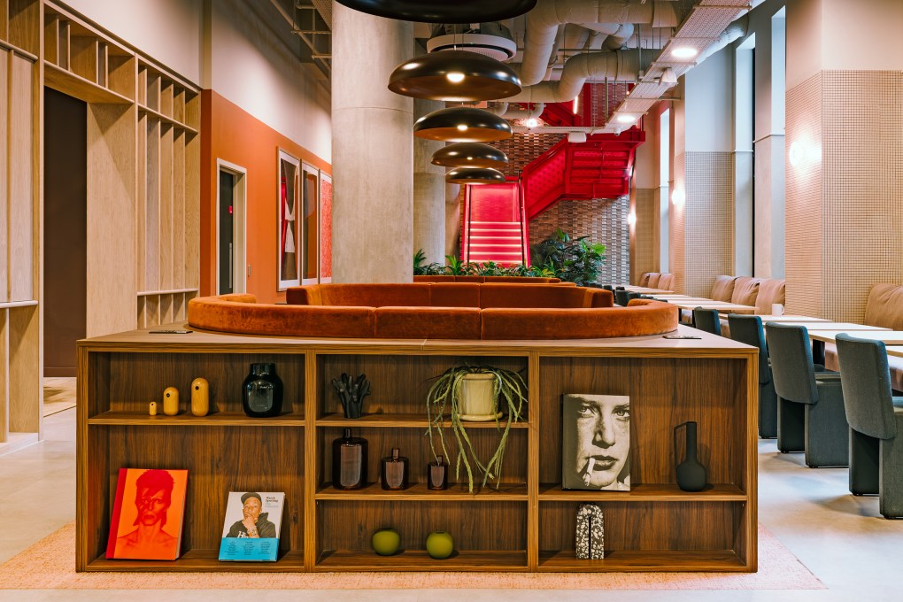 Modern lounge at Vita Student, with a wooden shelf displaying books, art, and decor. A circular orange sofa sits on top; red stairs and pendant lights are in the background. Tables and chairs line the right side of the space.