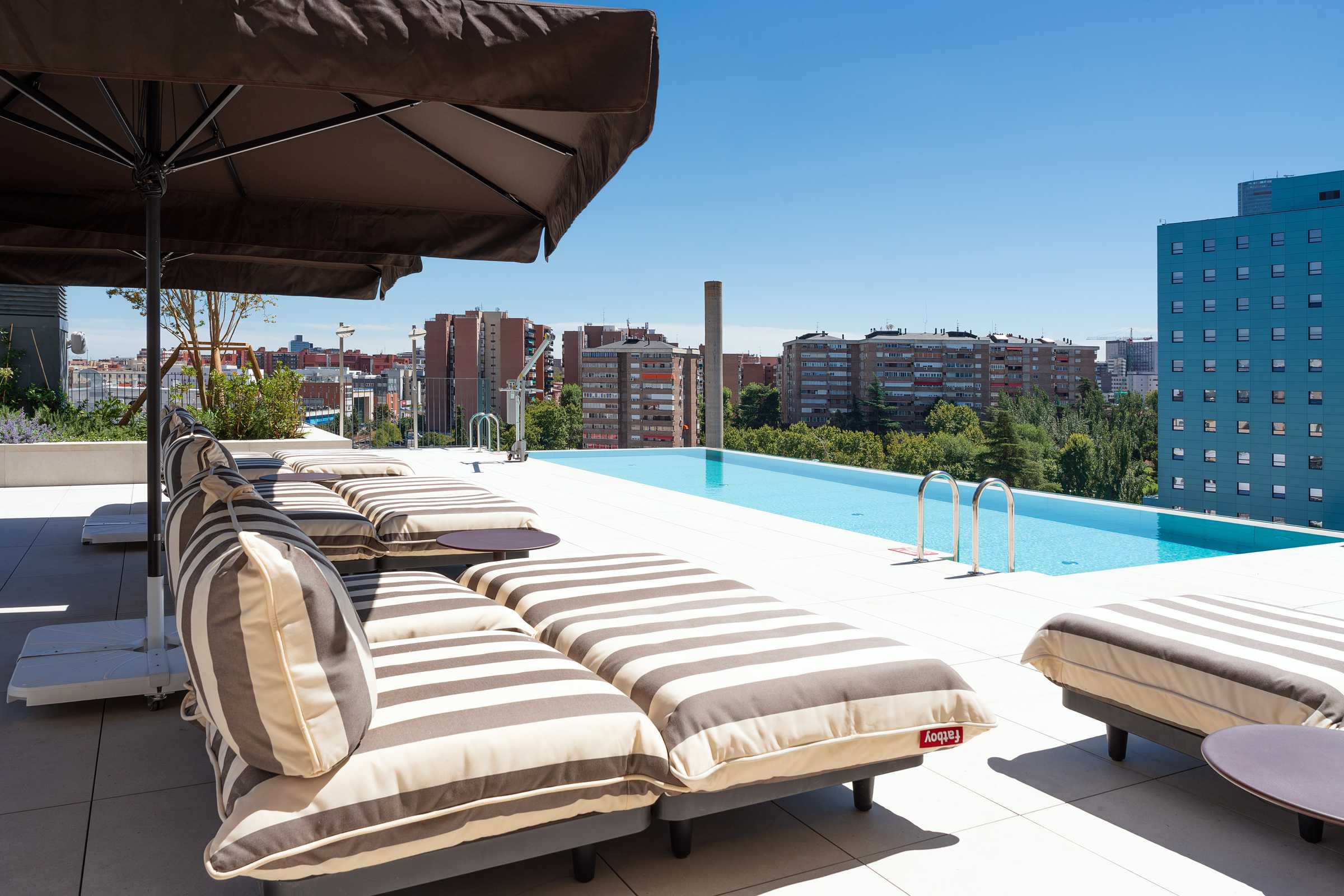 Striped lounge chairs sit beside a rooftop infinity pool under a large umbrella, with a city skyline and blue sky in the background.