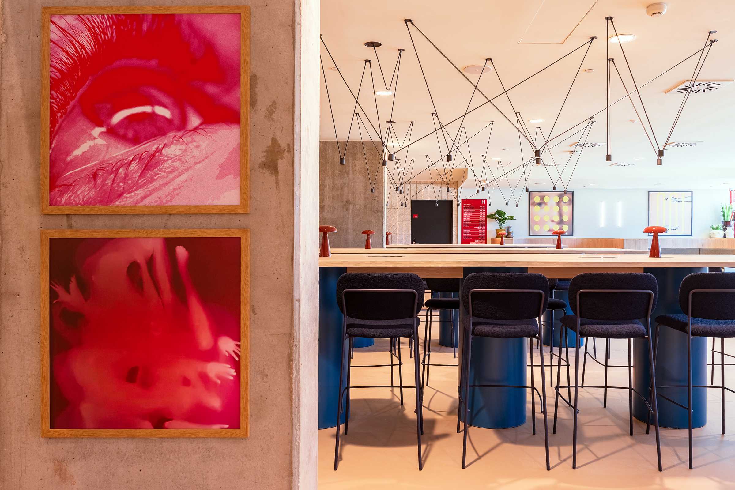 Two framed abstract red artworks hang on a concrete wall next to a modern cafe area with high tables, dark chairs, and overhead black wire light fixtures. The space is bright and contemporary.