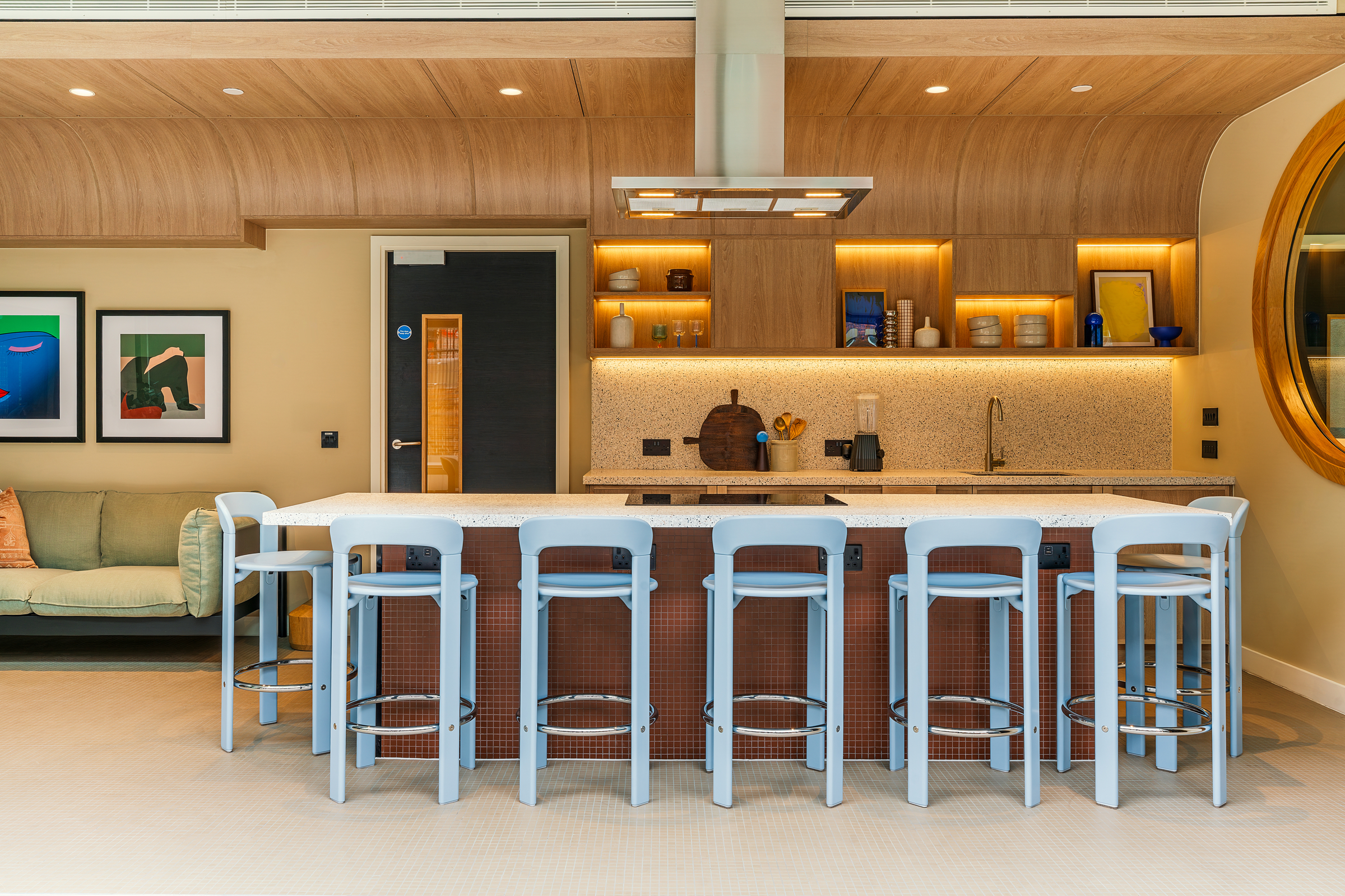 Modern kitchen with a marble island, six light blue bar stools, wood cabinets, open shelves with decor, and a round wall mirror. A green sofa and framed art are visible on the left side of the room, creating an inviting atmosphere perfect for an open day gathering.