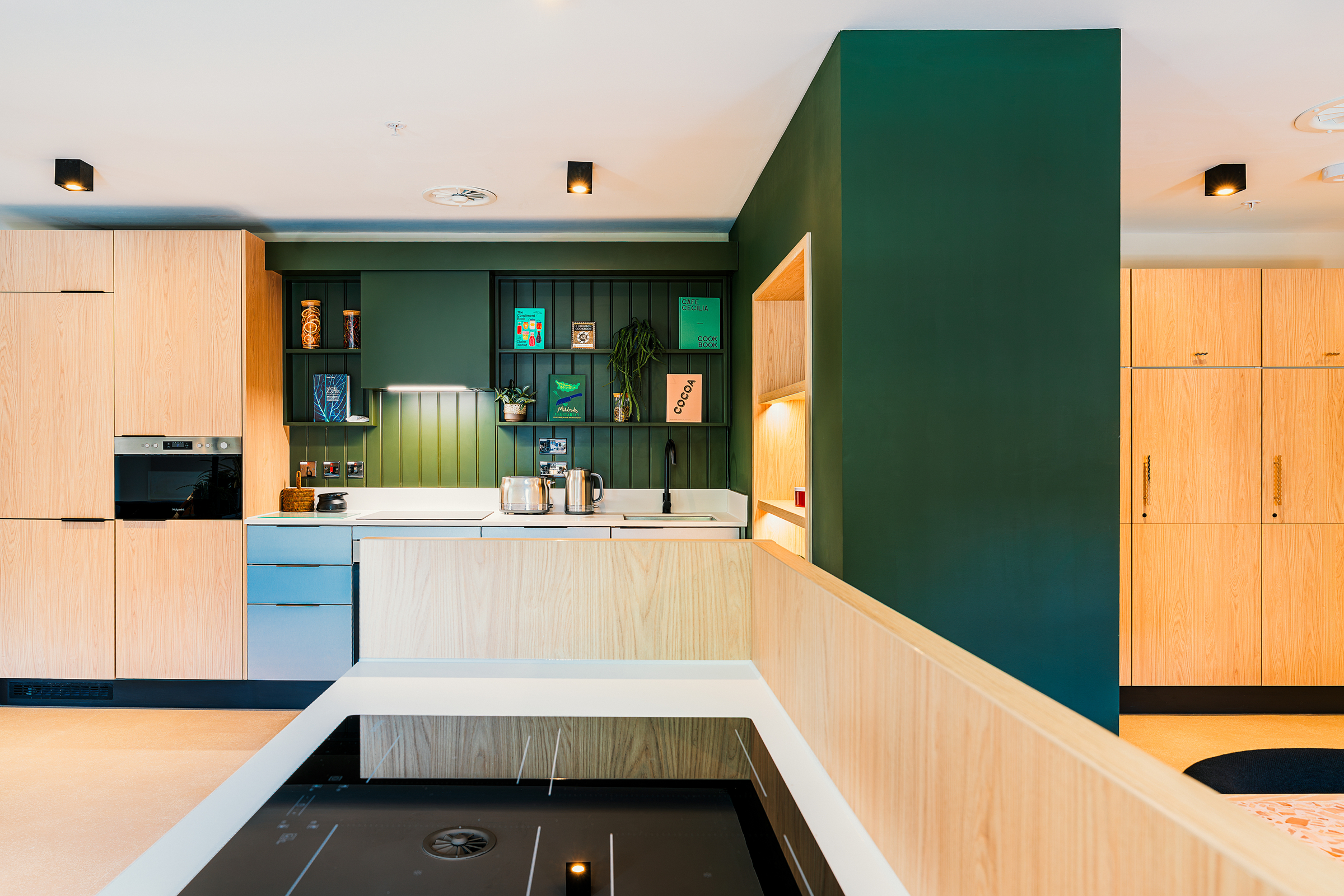 Modern kitchen with light wood cabinets, green accent wall, built-in appliances, white countertops, and open shelves with decor items—just the kind of bright and clean space you’d expect from Vita Student. The electric stove is in the foreground, enhancing the functionality and contemporary style of this inviting kitchen.