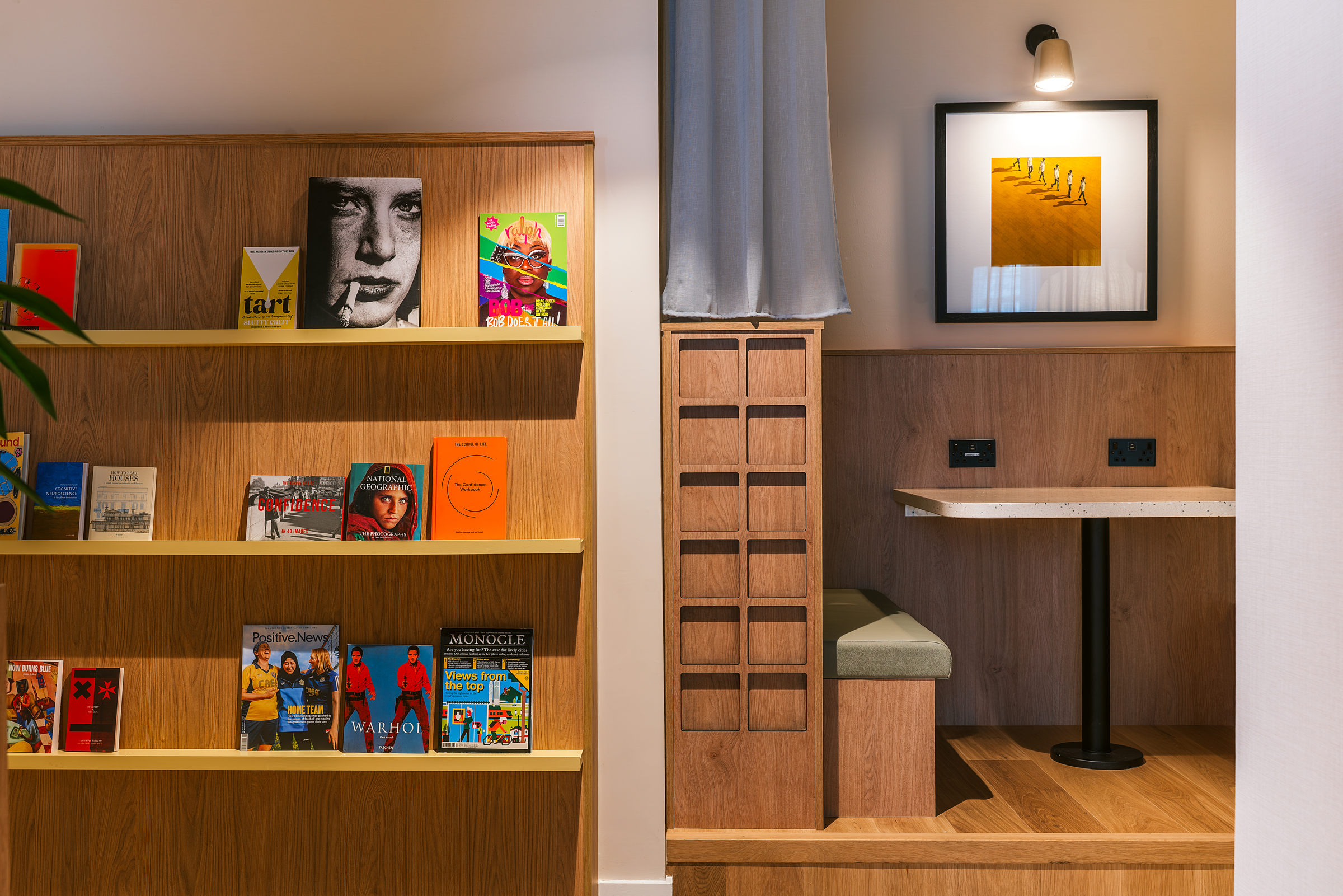 Wooden bookshelves with colorful books on the left; on the right, a small wooden booth with a table, cushioned bench, framed photo on the wall, and overhead light—an inviting setting perfect for relaxing or catching up during an open day.