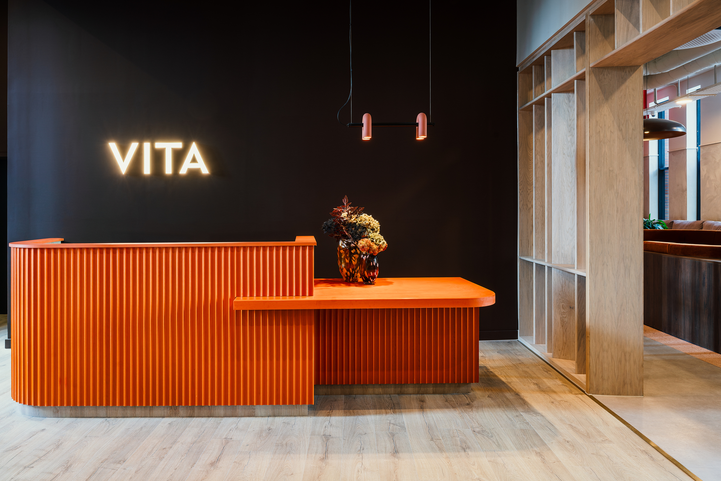 A modern Vita Student reception area with an orange desk, flower arrangement, two hanging lights, and a neon “VITA” sign on a black wall. Light wood flooring and an open wooden frame partition are visible.