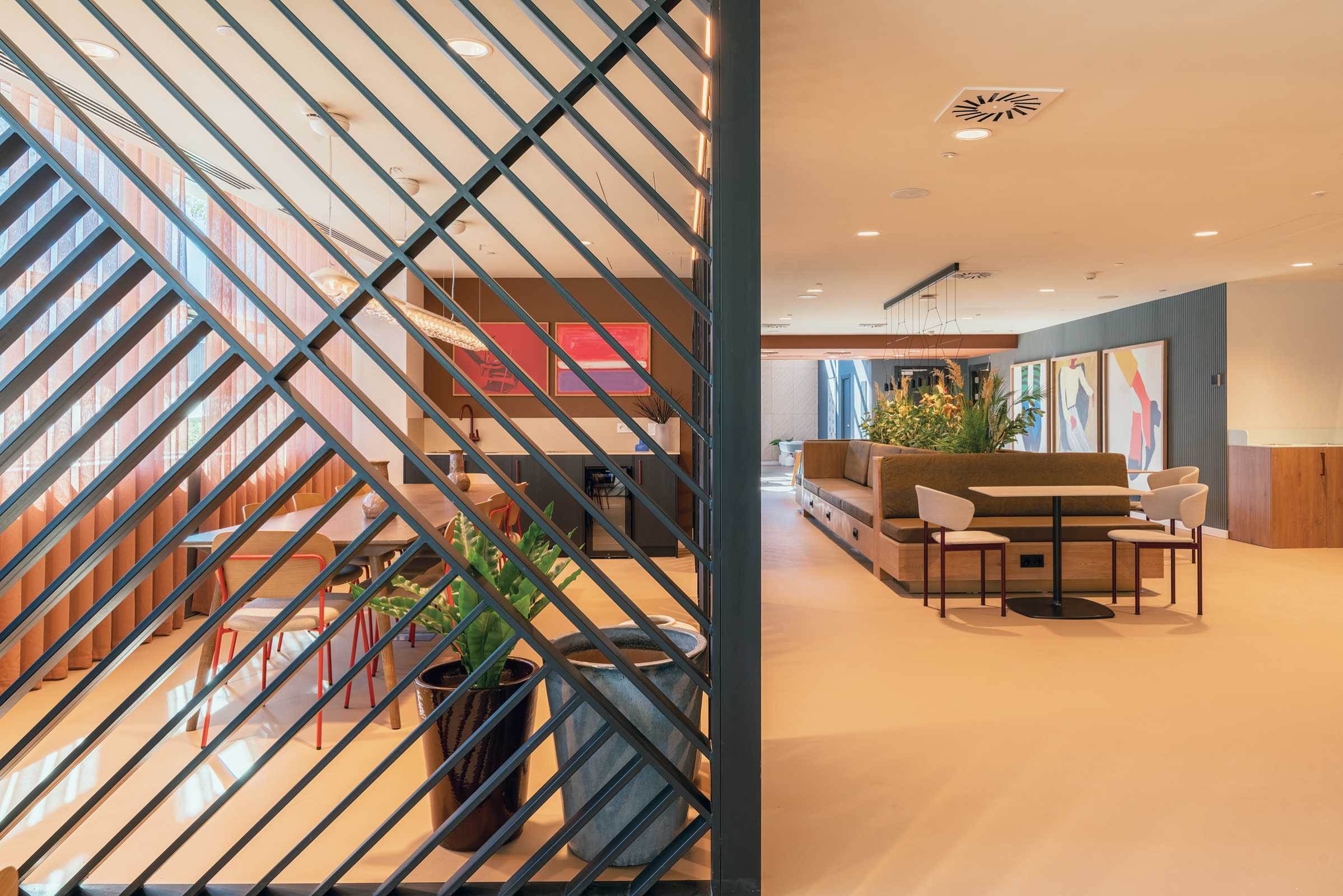 Modern office lounge with contemporary furniture, potted plants, abstract art on the walls, and a black slatted divider in the foreground separating two seating areas. Warm lighting creates an inviting atmosphere.