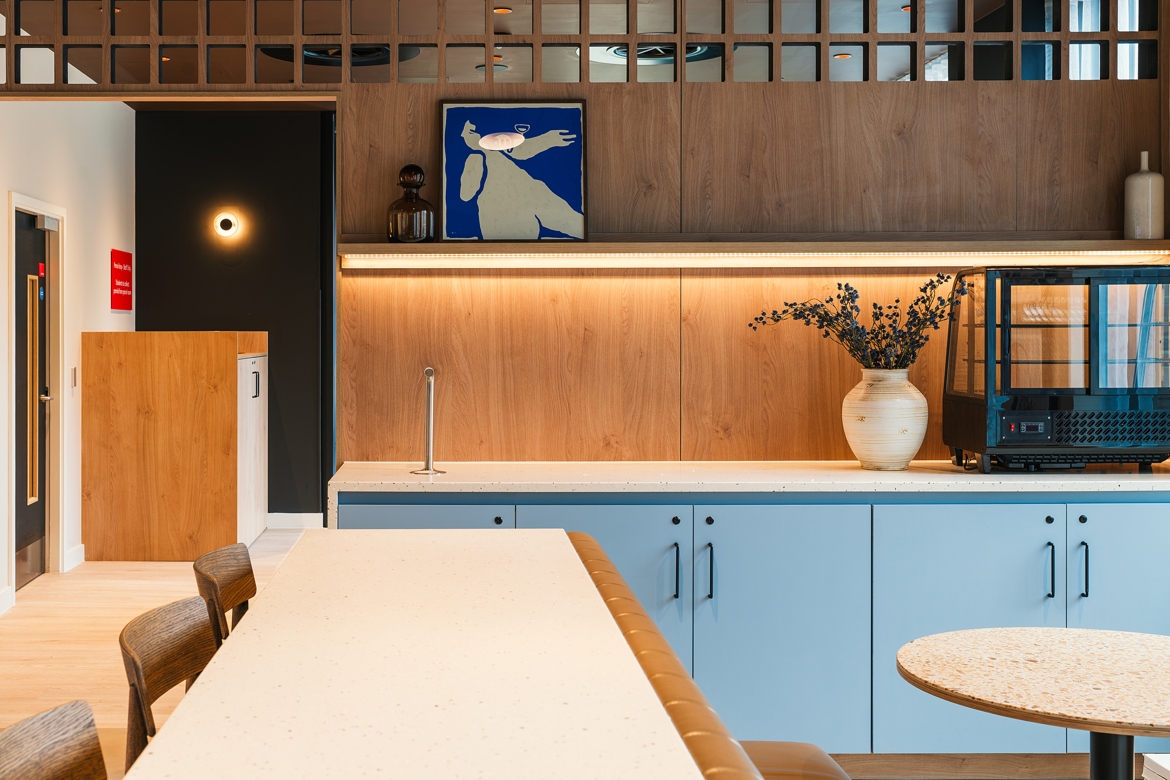 Modern kitchen with light blue cabinets, a beige countertop, wooden chairs, a large vase with flowers, wall art, and warm lighting under wooden paneling. A black wire frame and decor items sit on the counter, creating an inviting atmosphere perfect for an open day event.