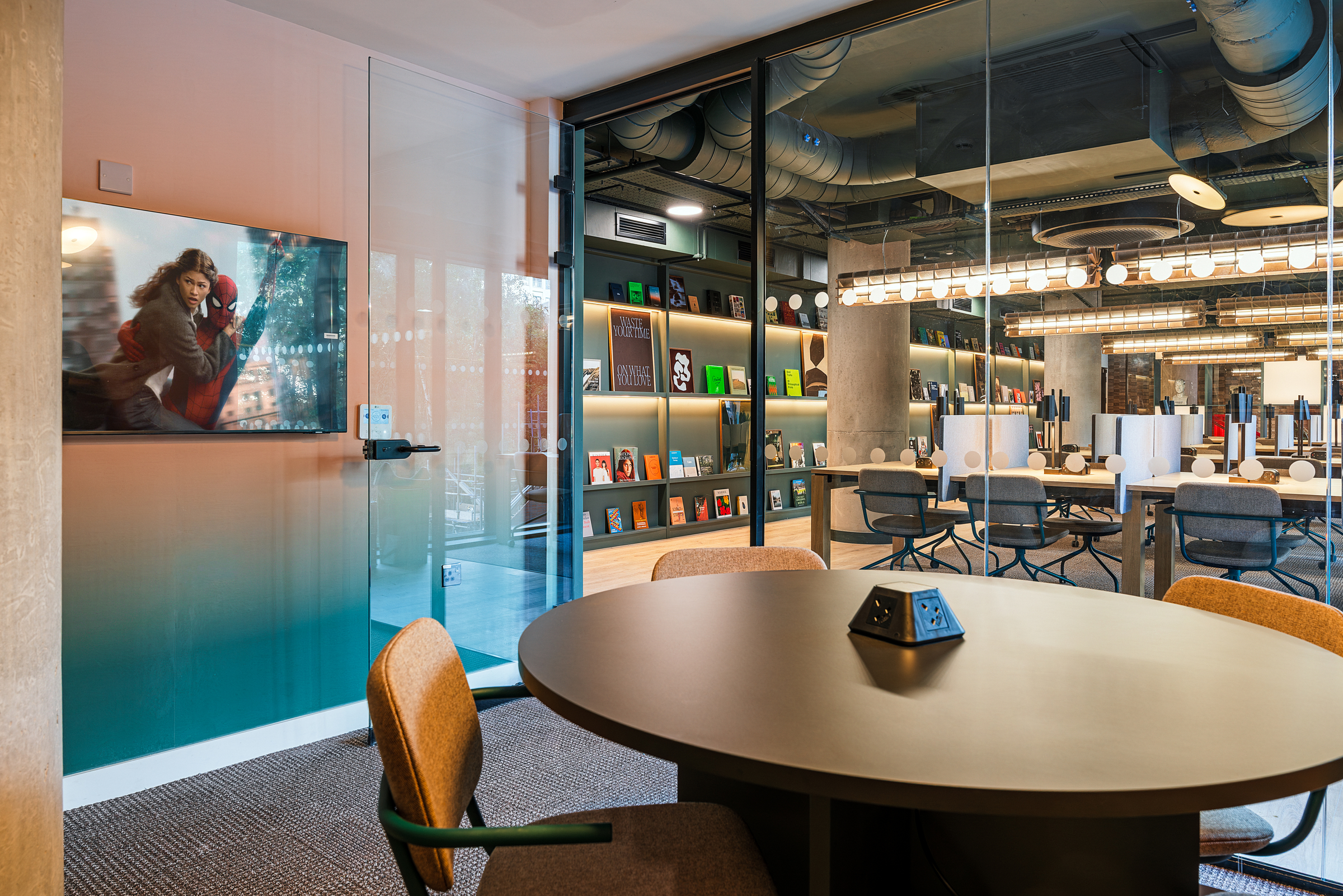 Modern office space with a round table and chairs in a small meeting room at Vita Student, glass walls, a bookshelf with books in the background, and a TV displaying a scene from a movie.