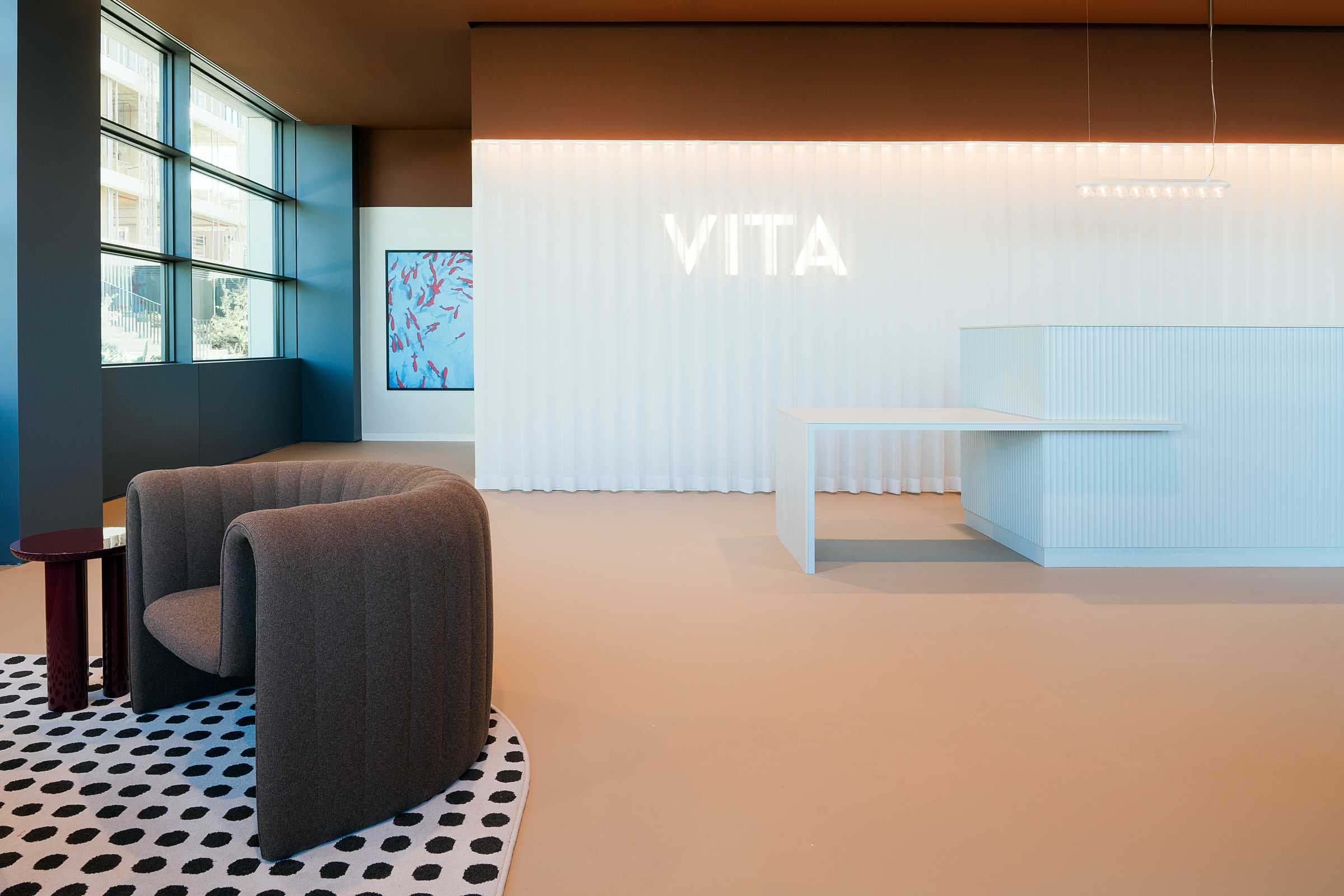 Modern office lobby with a rounded brown chair on a polka-dot rug, a white reception desk, wall art, and “VITA” illuminated on a white textured wall beside large windows.