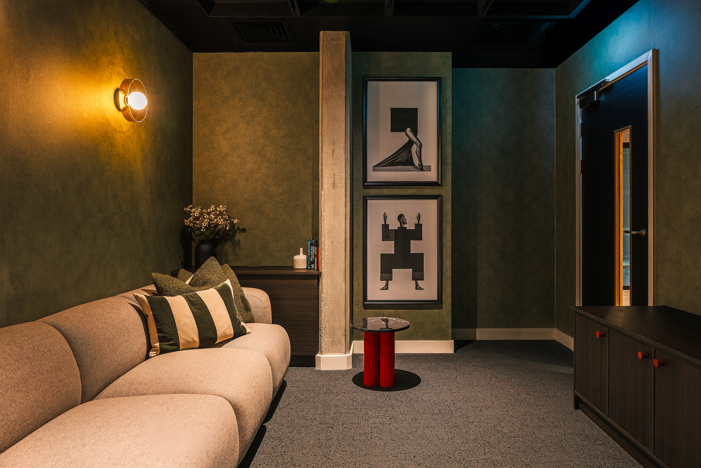 A cozy, modern lounge with a beige sofa, striped pillows, side table, and wall art. The room has green walls, dim lighting, and minimalistic decor, creating a warm and inviting atmosphere—perfect for relaxing or welcoming guests during an open day.