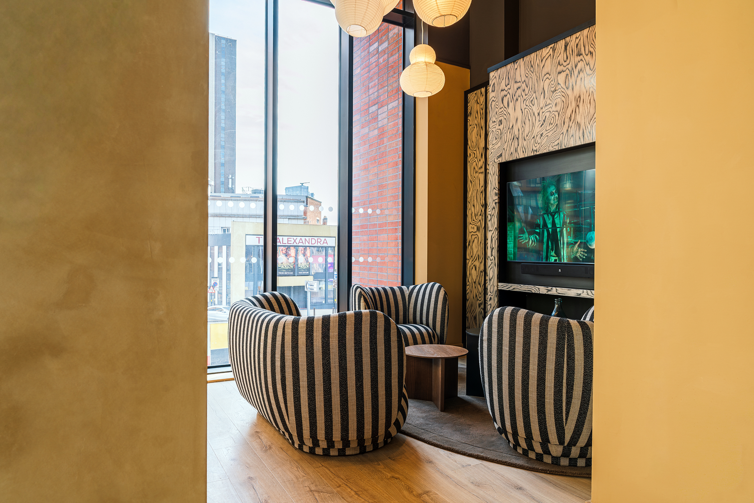 A cozy lounge area at Vita Student features three striped chairs, a small round table, pendant lights, a large window, and a TV displaying a movie scene. City buildings are visible outside the window.