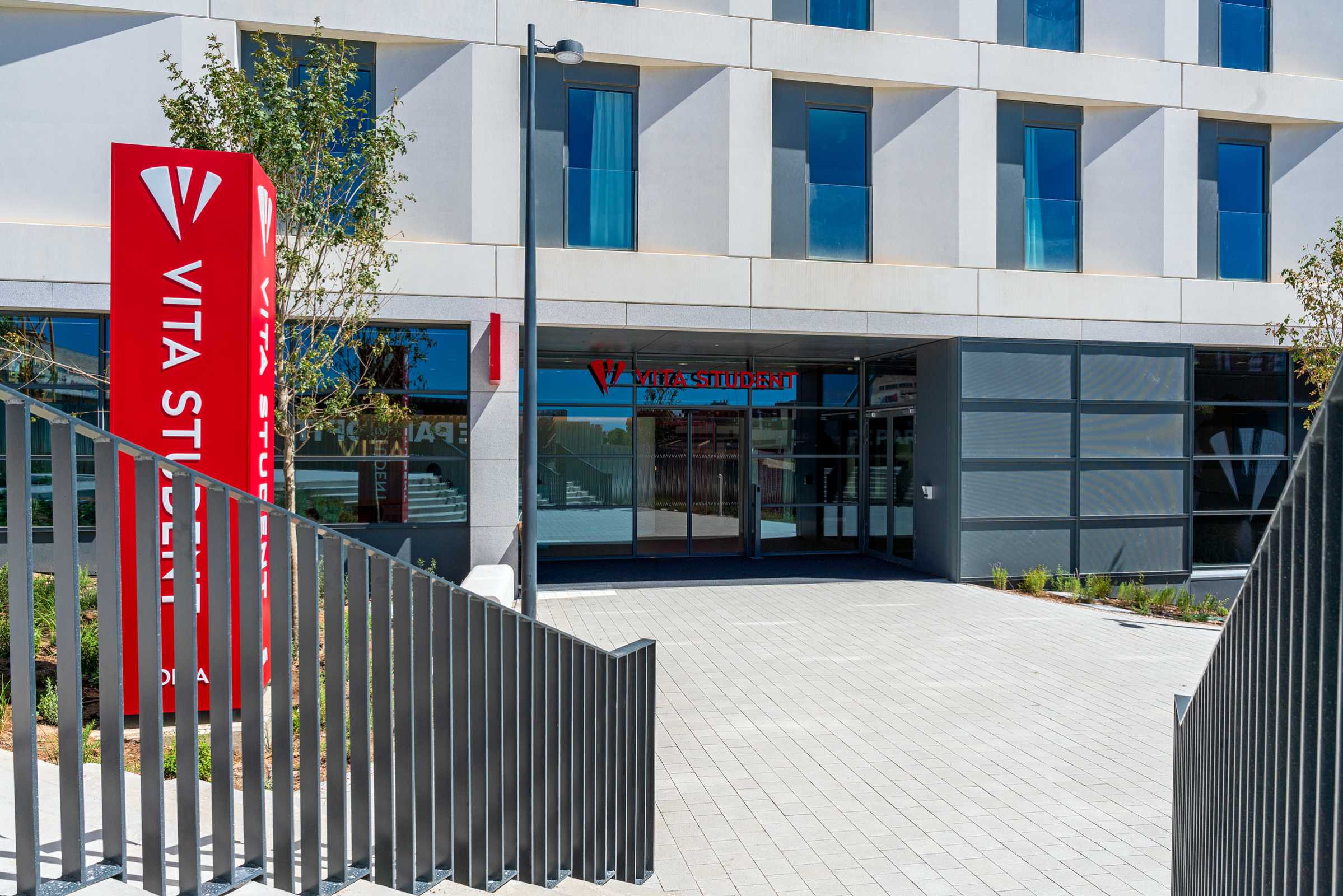 Modern student accommodation building with large windows, a red VITA STUDENT sign near the entrance, and a paved pathway bordered by metal railings leading to the front doors.