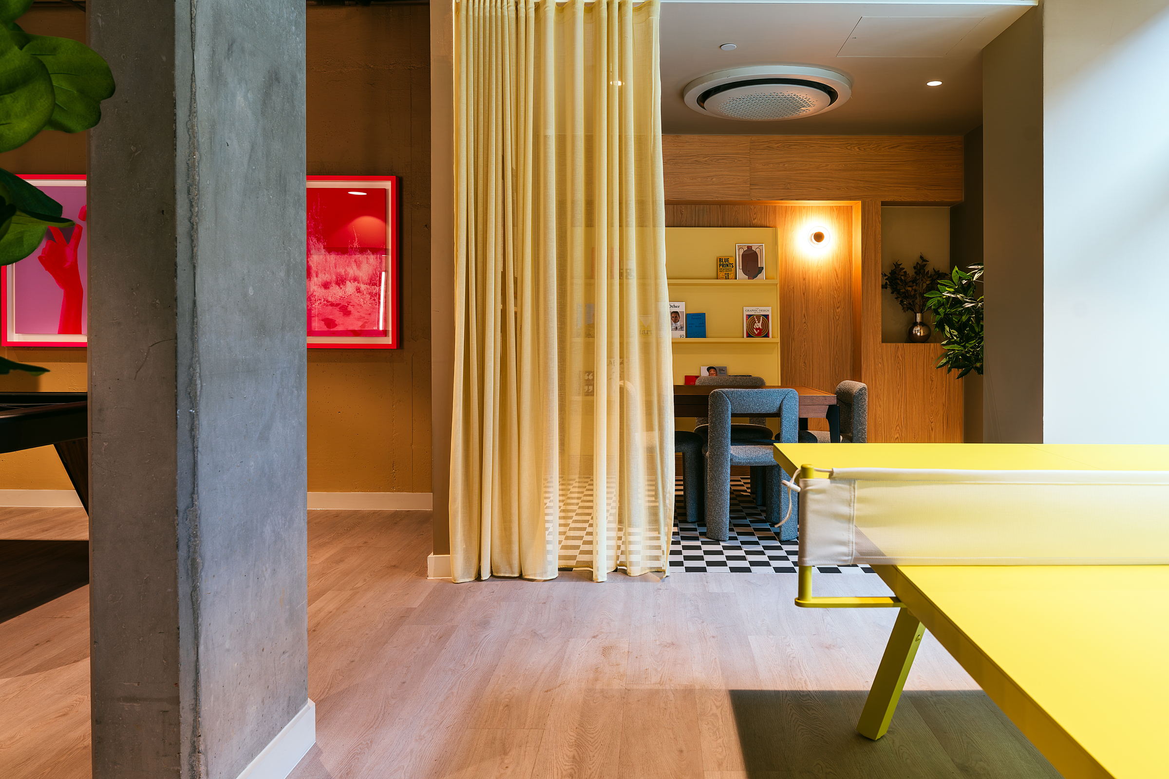 Modern indoor space with a yellow ping pong table, sheer yellow curtain, concrete pillar, framed art on the wall, and a cozy seating area with checkered floor in the background—perfect for hosting an open day.