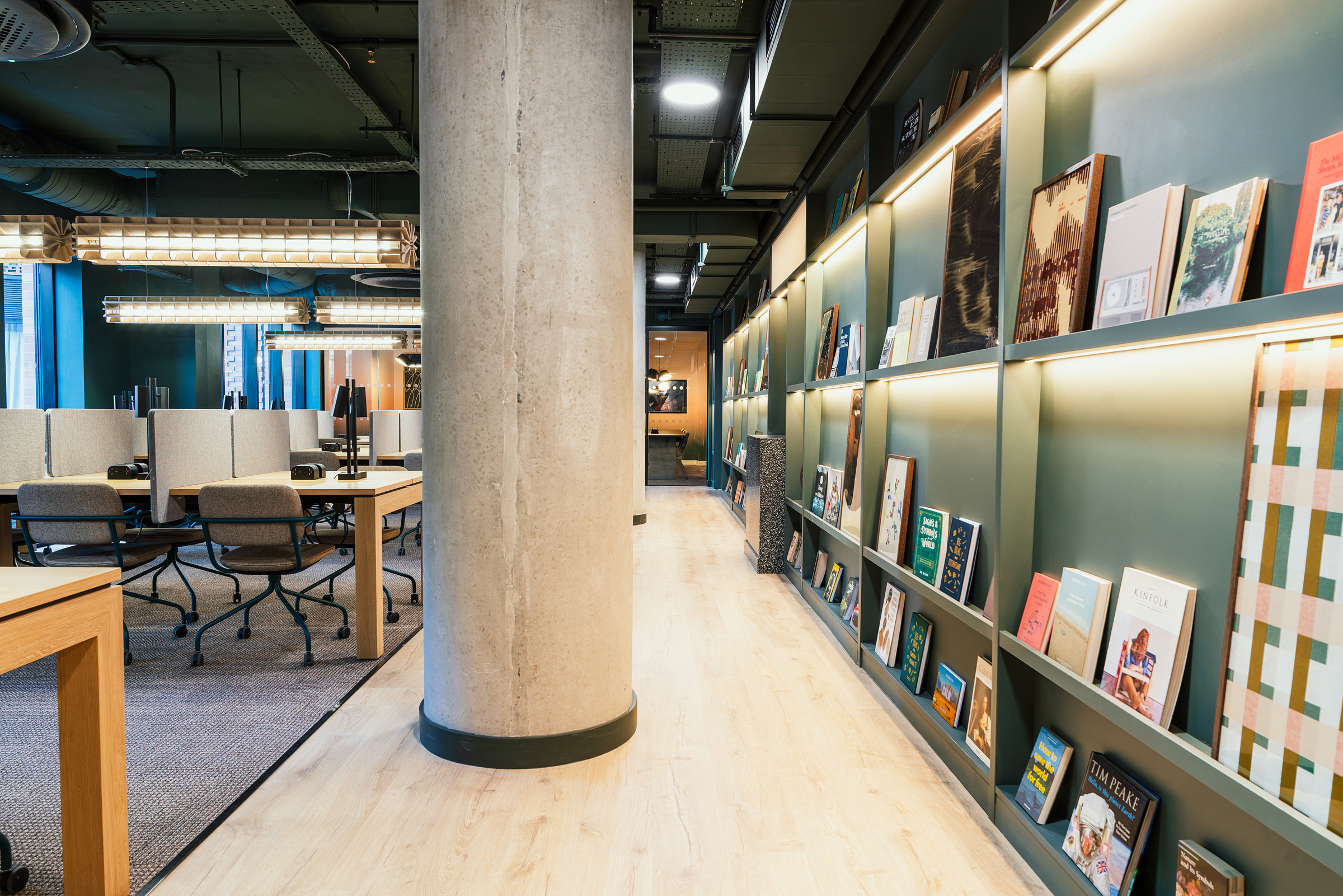 Modern Vita Student office space with open desks and chairs on the left, and a hallway with books displayed on shelves to the right. A large concrete pillar stands in the center. The area is brightly lit and organized.