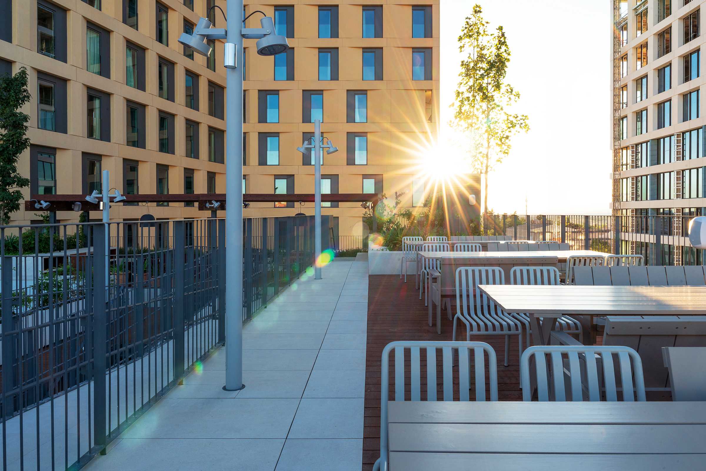 Outdoor patio with empty white tables and chairs beside a metal fence. Modern apartment buildings surround the area. The sun is shining brightly between the buildings, casting a lens flare over the scene.