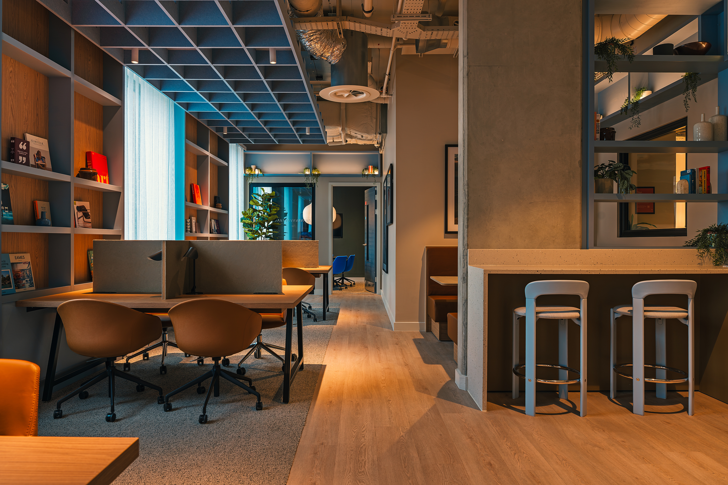 Modern coworking office with desks, brown chairs, bookshelves, high stools at a counter, and cozy lighting. Plants and decor create a warm, inviting atmosphere with a mix of open and private workspaces.