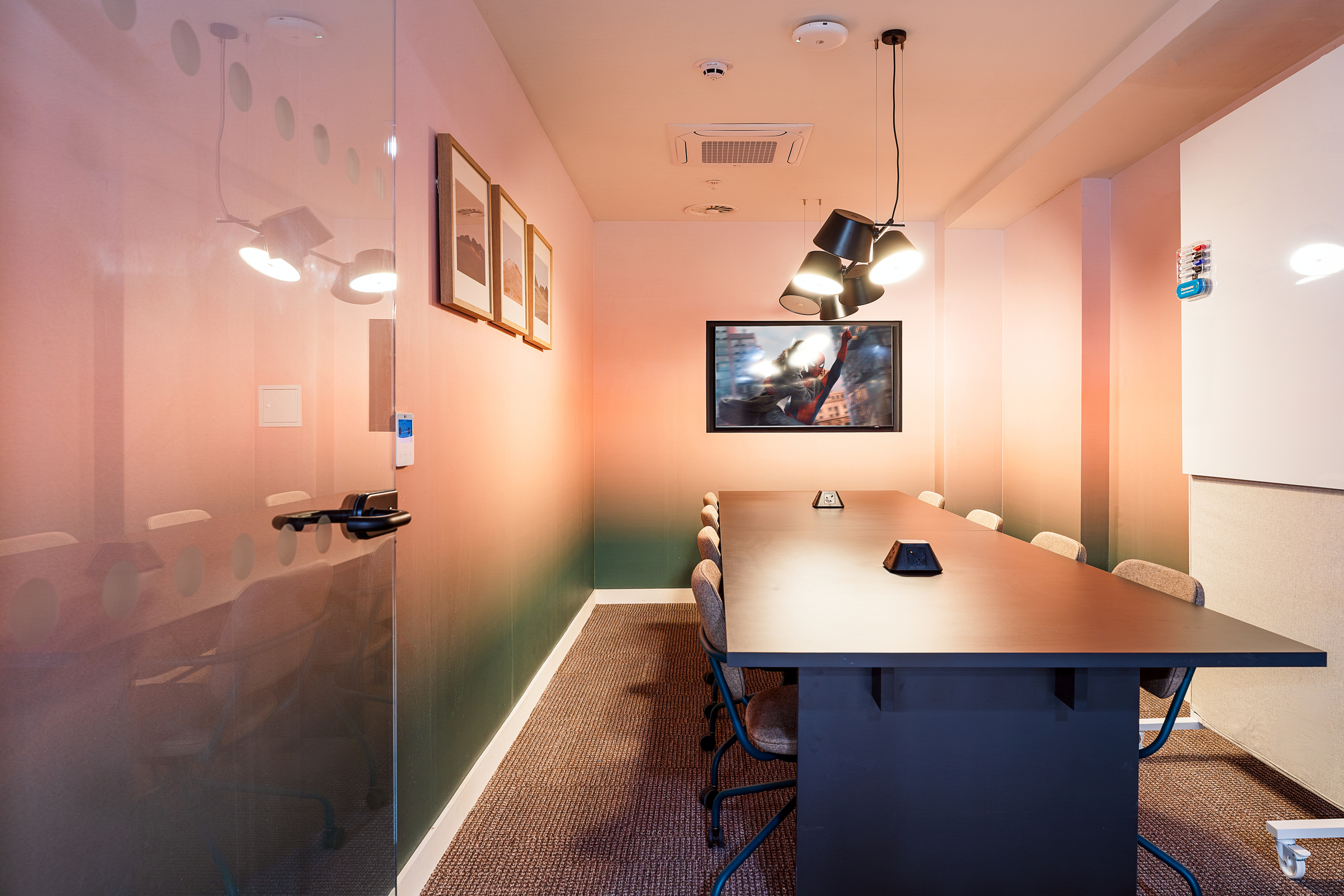 A modern Vita Student conference room with a long table, eight chairs, wall art, ceiling lights, and glass door. The room features gradient-colored walls and a mounted screen at the far end.