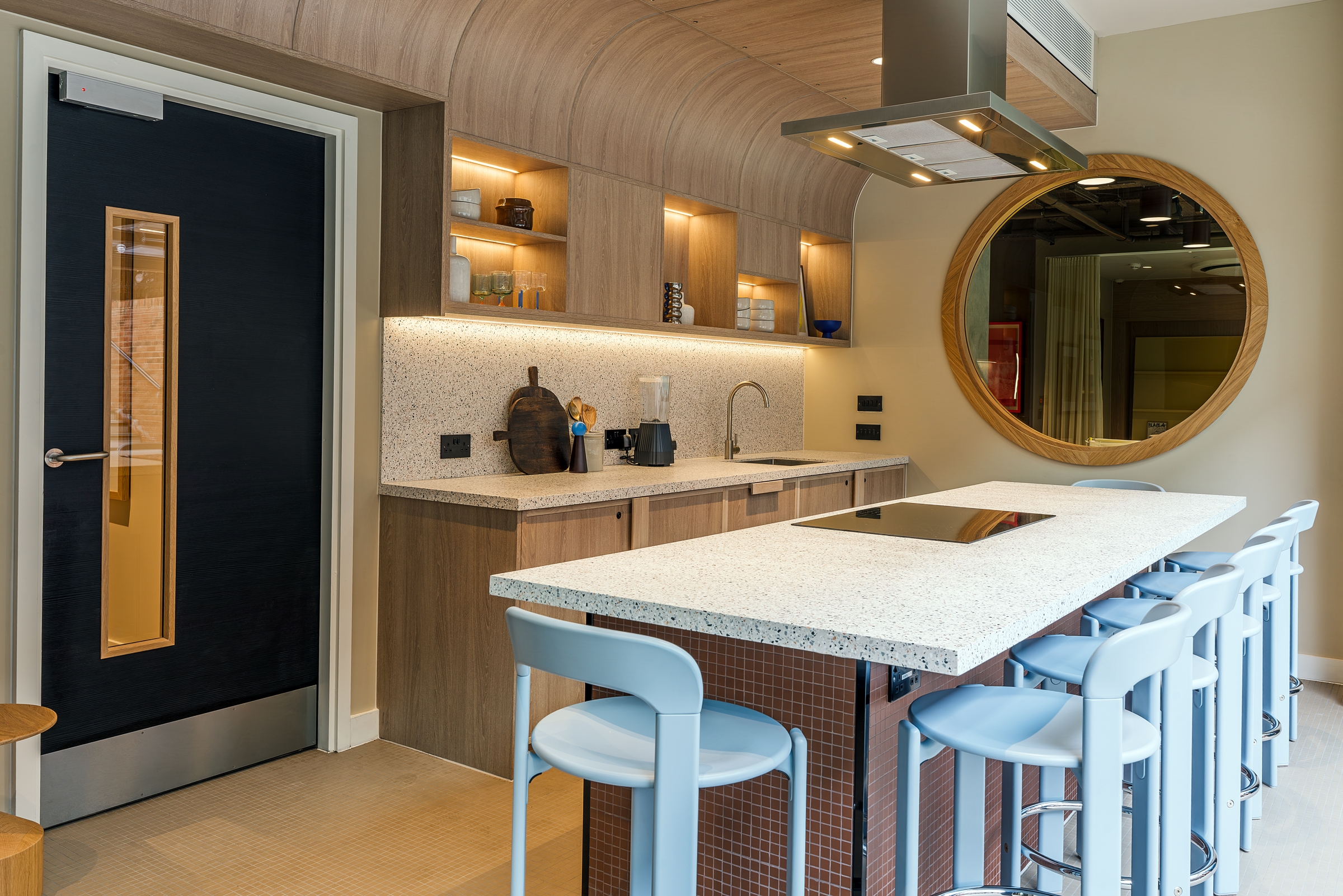 Modern kitchen with wood cabinets, open shelves, a large round mirror, induction cooktop, and four light blue bar stools at a speckled countertop island. Warm lighting and neutral tones create a cozy atmosphere.