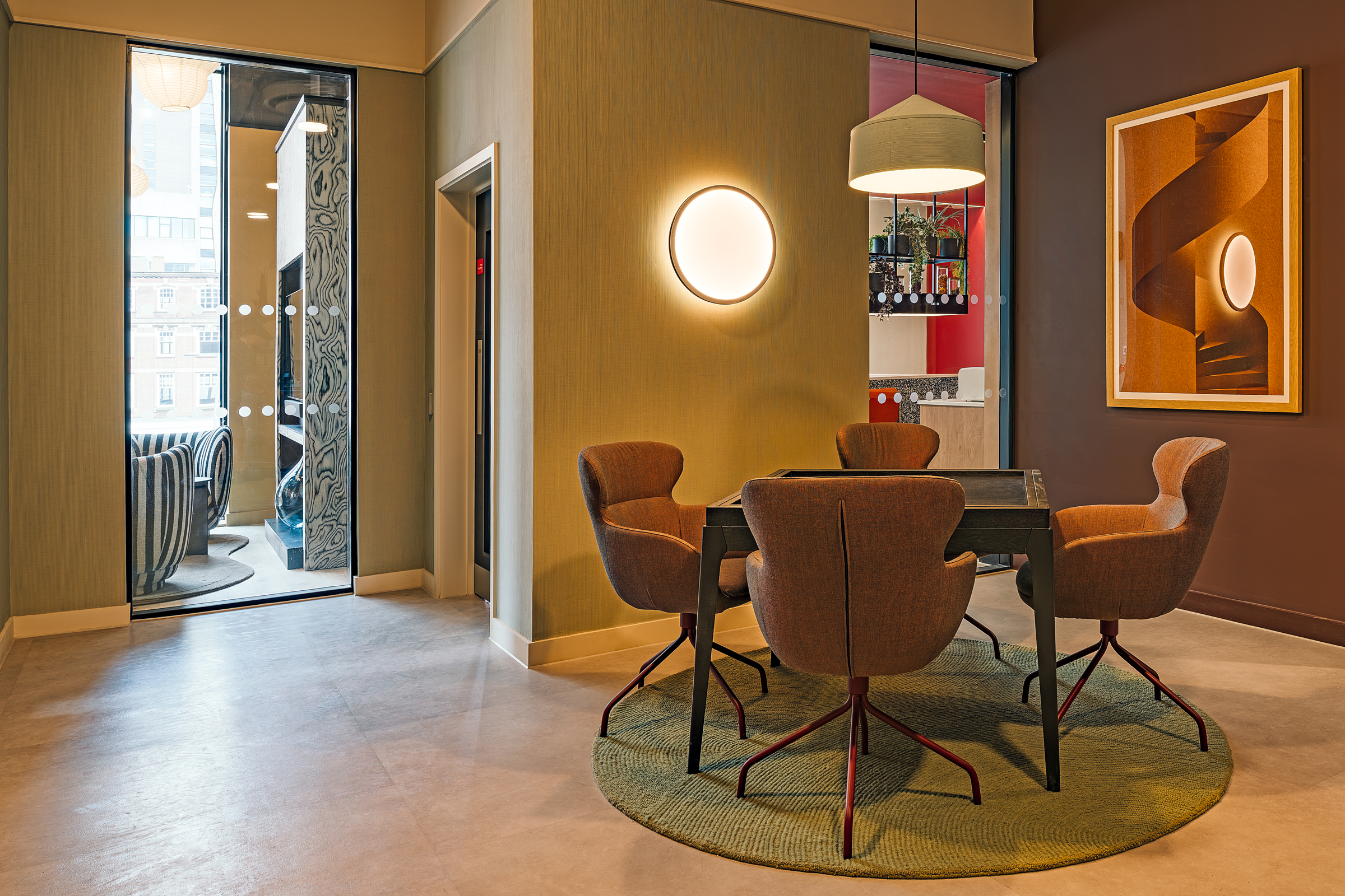 Modern meeting room with four brown chairs around a dark square table on a green rug, circular wall light, framed abstract art, and glass door showing another room with chairs and windows.