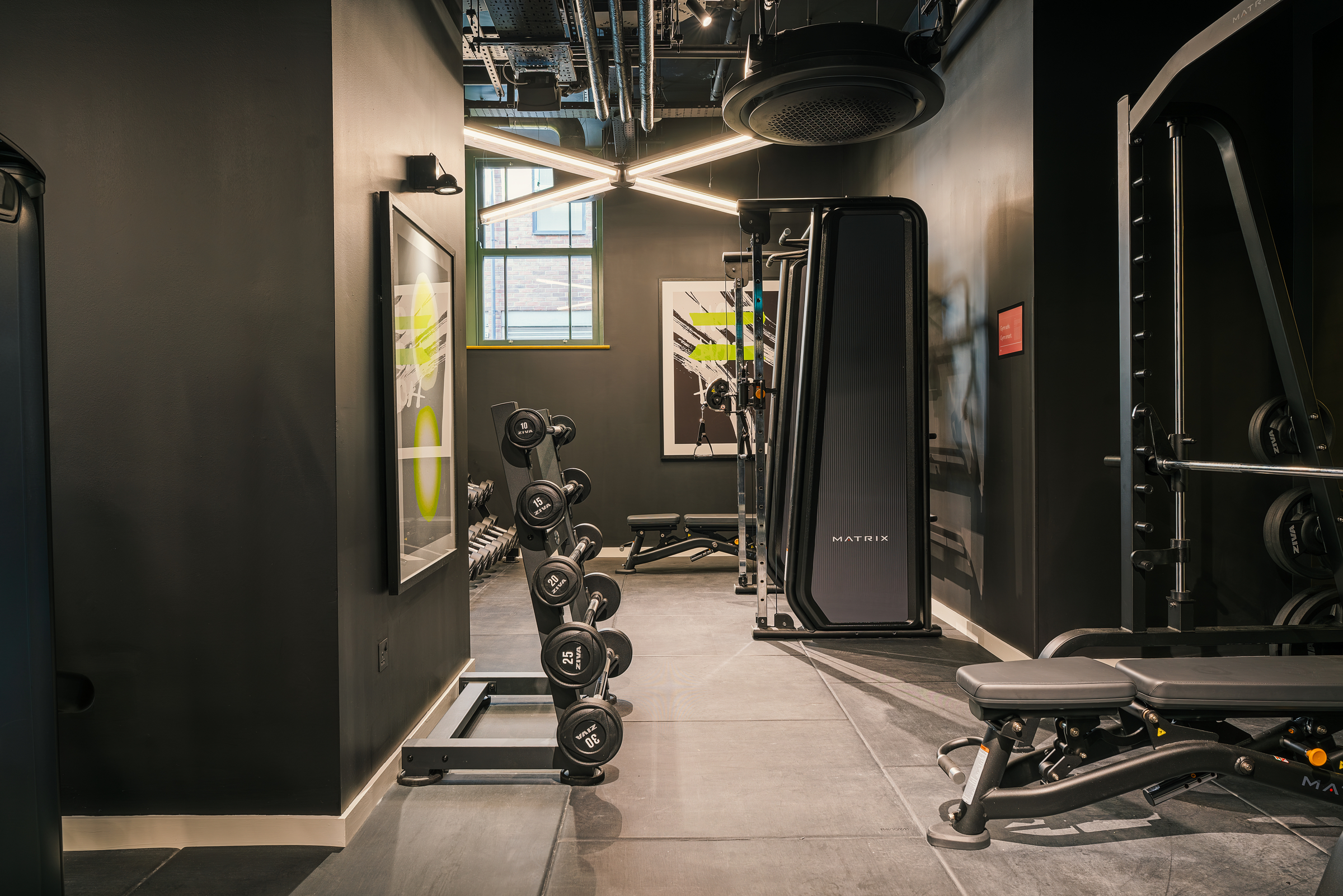 A modern gym with free weights, weight machines, a bench, and fitness artwork on the walls. The space is well-lit with industrial-style lighting and a window letting in natural light.