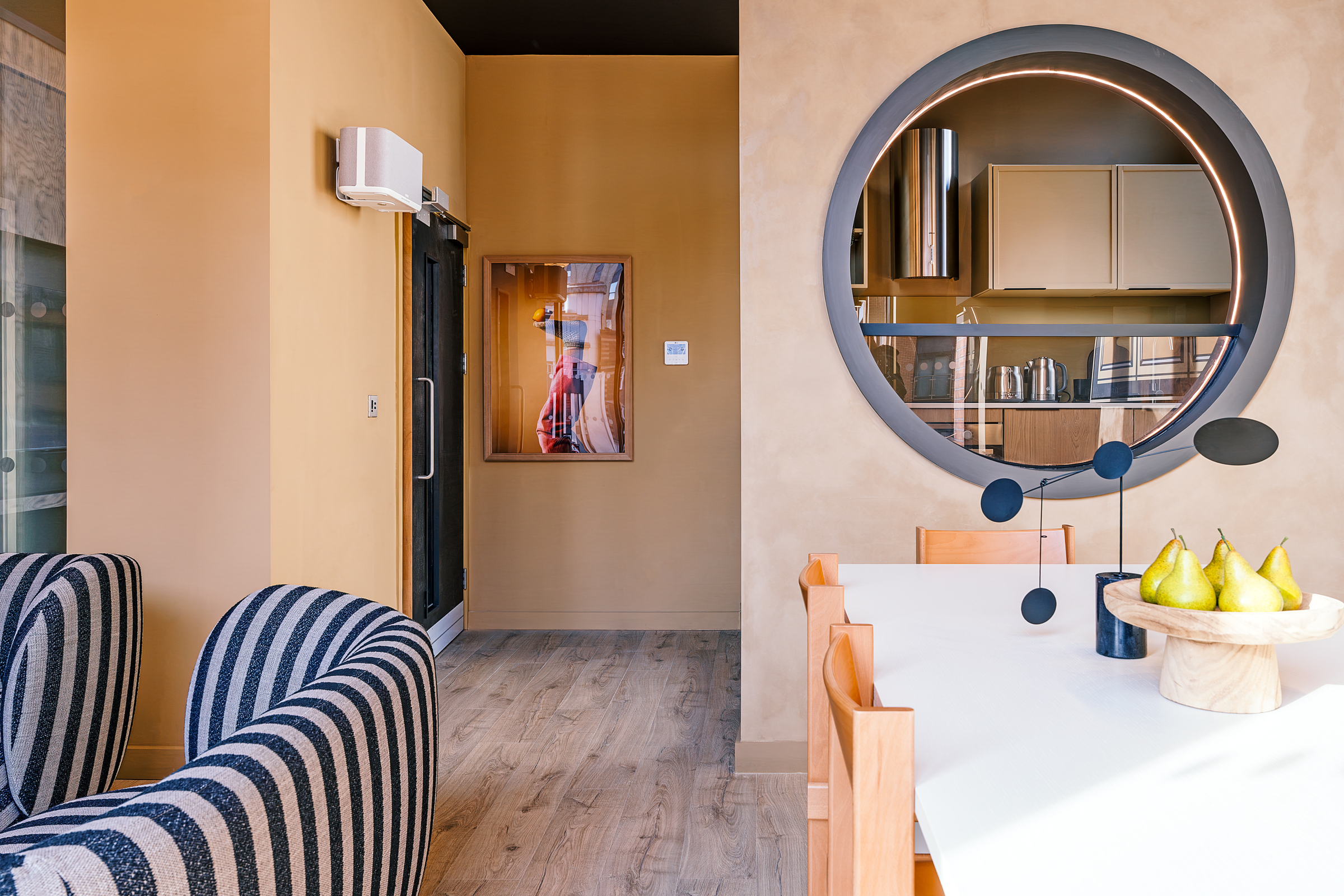 Modern dining and living area with a white table, wooden chairs, black abstract centerpiece, bowl of pears, striped armchairs, and a circular mirror reflecting a kitchen. Warm beige walls and wood flooring create a cozy atmosphere.