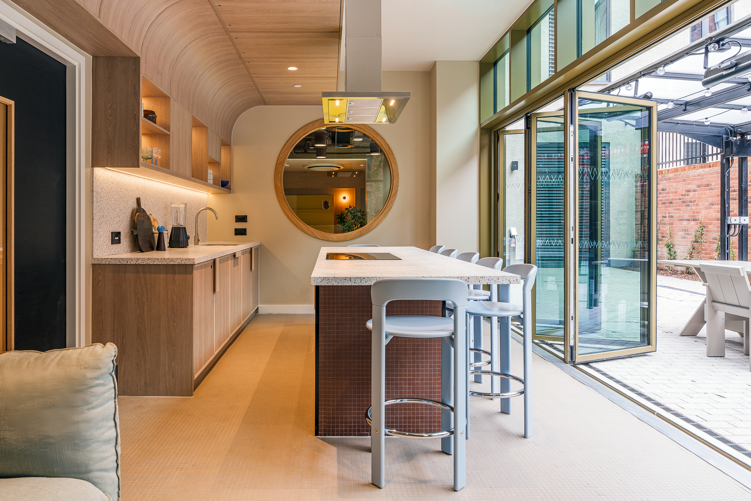 Modern kitchen with wood accents, a large round mirror, an island with three gray barstools, open shelving, and large glass doors opening to a patio with outdoor seating. Natural light fills the stylish, tidy space.