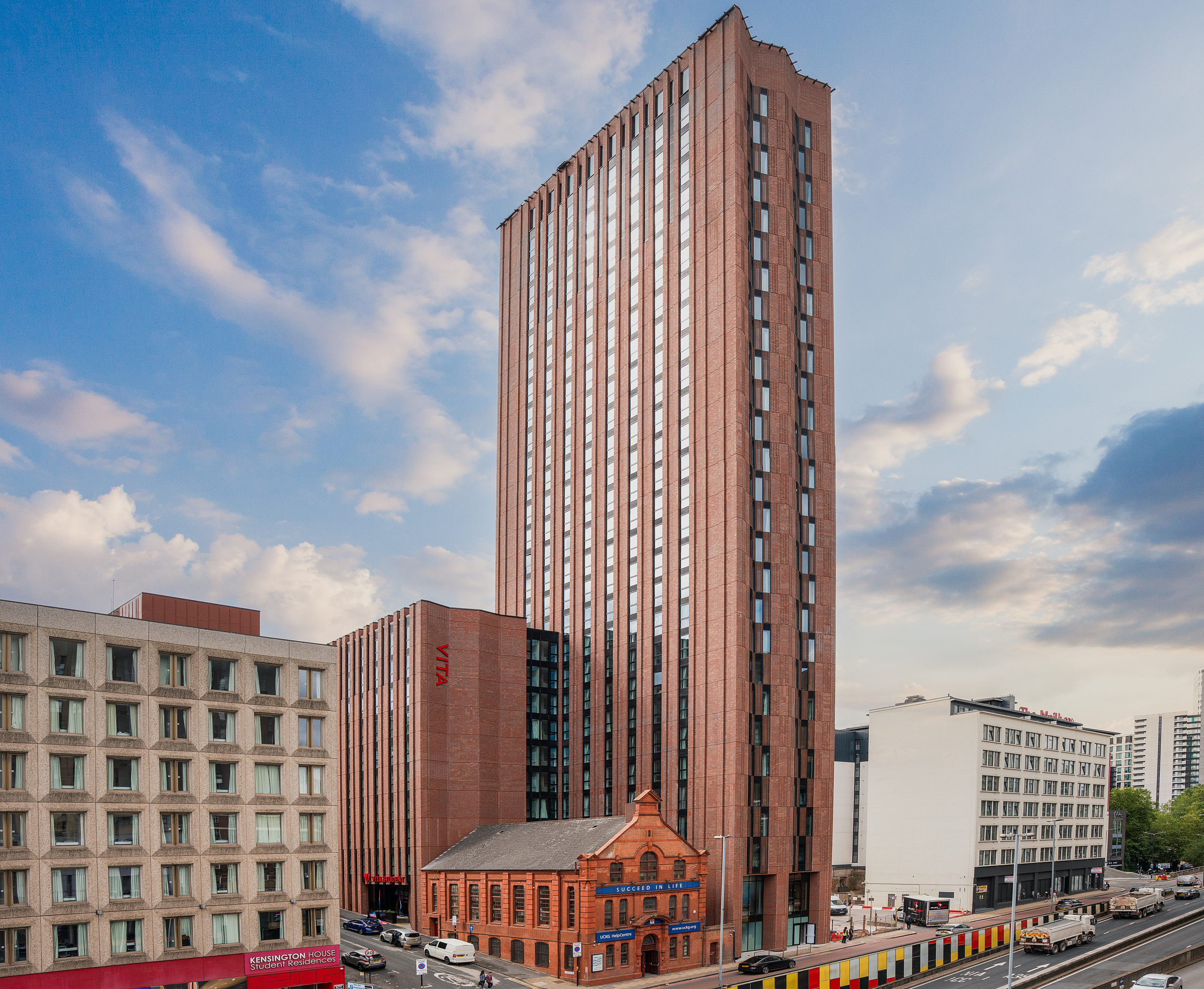 A tall, modern red-brick tower rises above an older, smaller red-brick heritage building. To the left is a concrete student residence, and in the foreground, a road with traffic barriers, all set against a blue sky with soft, scattered clouds.