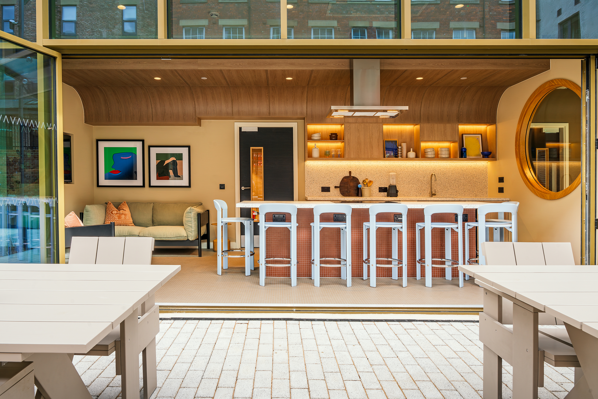 Modern open-plan kitchen and lounge with a curved wooden ceiling, bar stools at a counter, framed artworks on the wall, a round mirror, and white patio tables in the foreground, seen through folding glass doors.