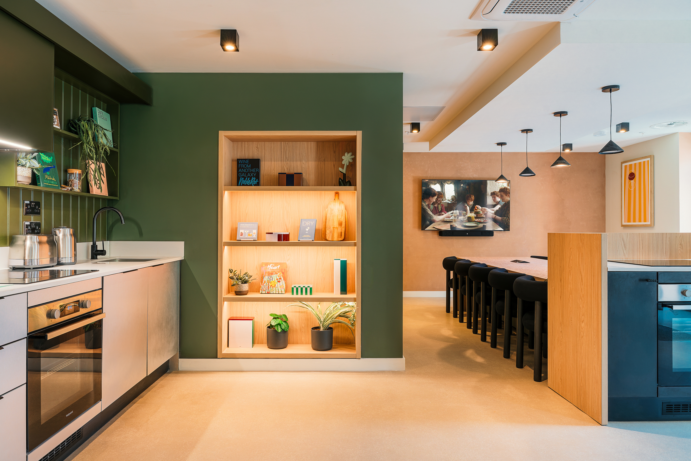 Modern kitchen with green and beige walls, open shelving displaying books and plants, a long dining table with black chairs, and pendant lights hanging from the ceiling—perfectly embodying the stylish, communal atmosphere Vita Student is known for.