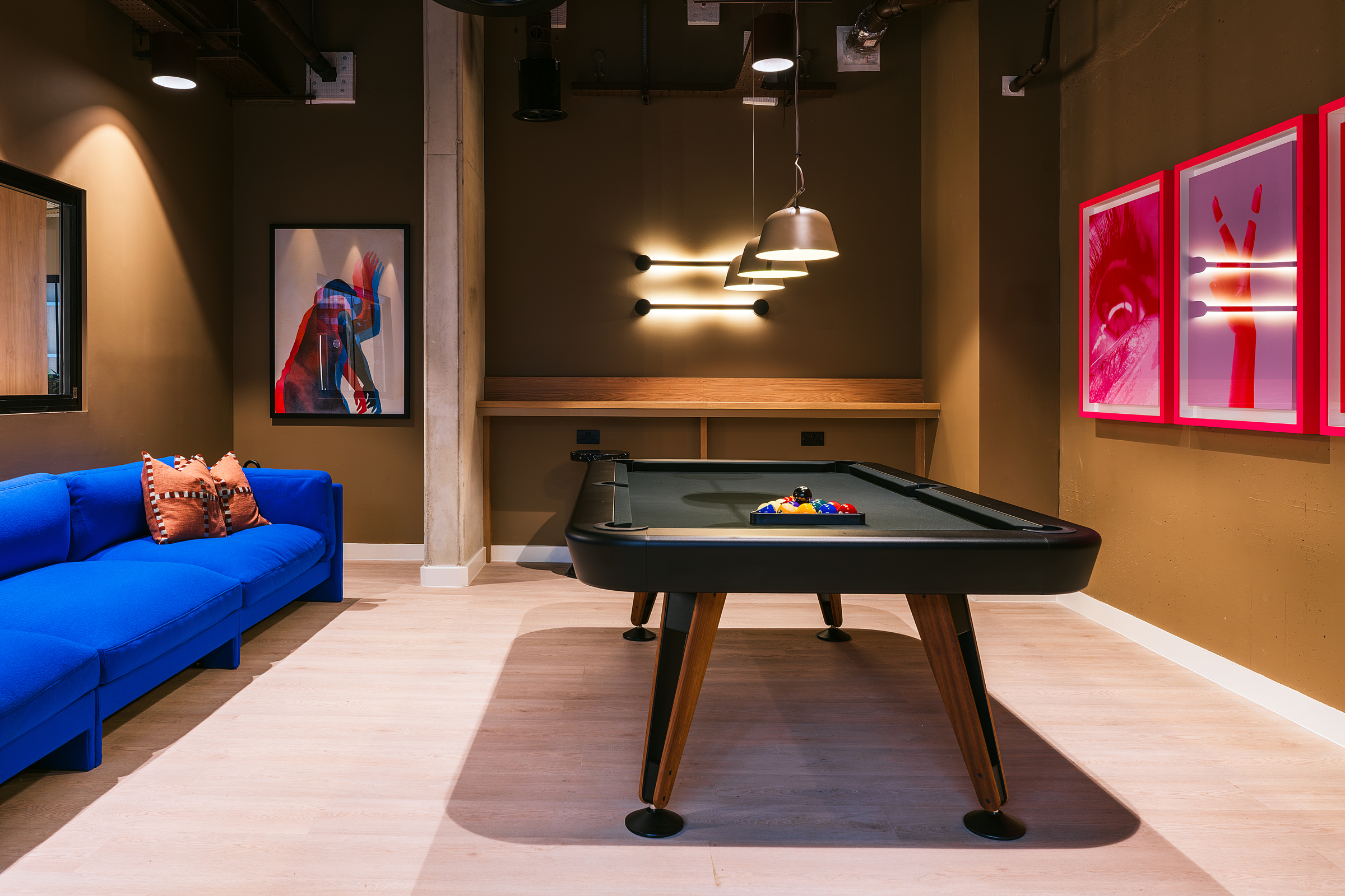A modern game room with a black pool table, blue couch, colorful artwork on brown walls, and two pendant lights above the table welcomes visitors on open day. Pool balls and cues are set up for play, inviting everyone to enjoy the vibrant space.