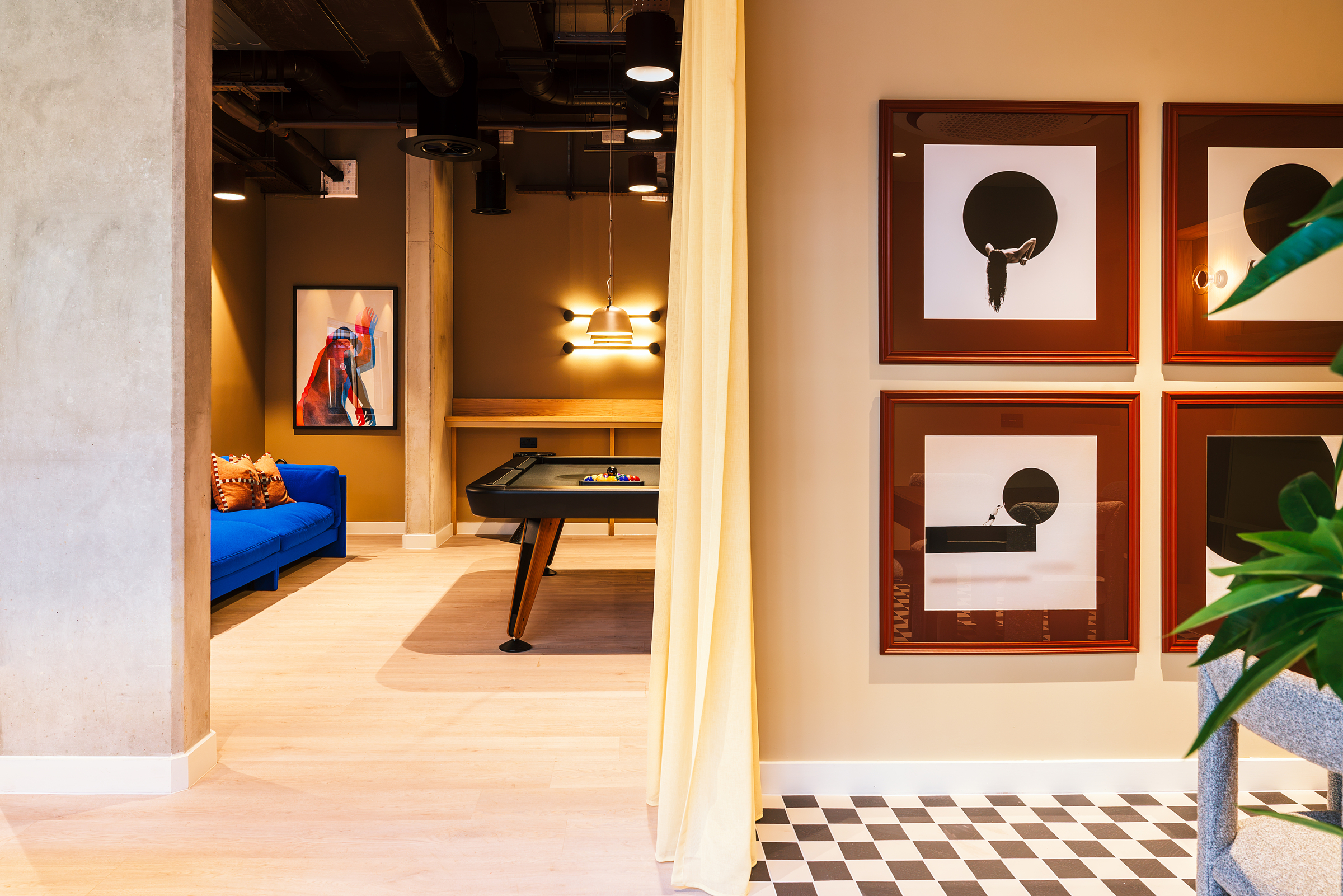A modern lounge with a blue sofa, a pool table, framed abstract art on tan walls, and a checkered floor section. A yellow curtain partially divides the space; green plants are in the foreground.